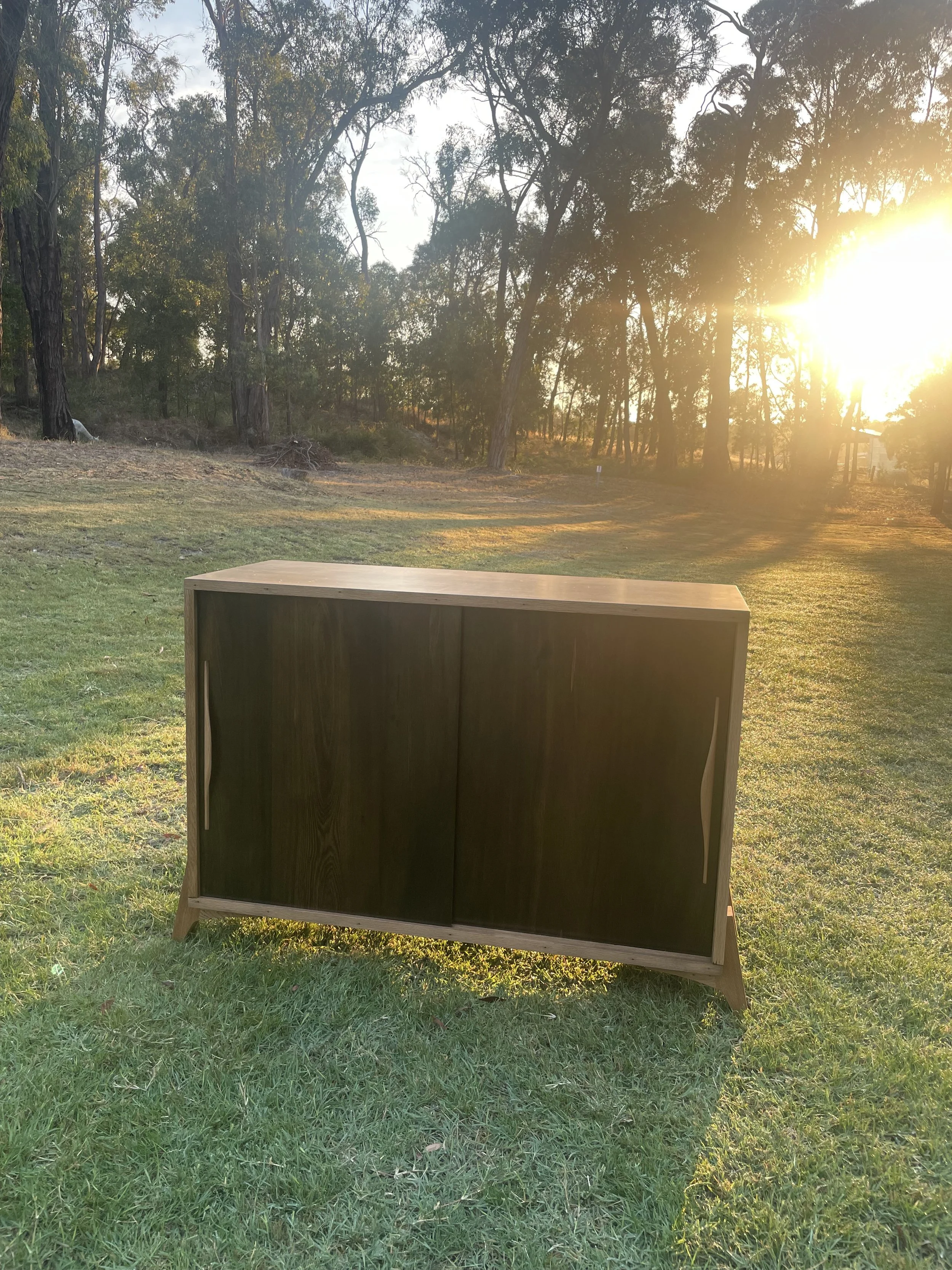 A wooden cabinet with sliding dark doors outdoors on a grassy field during sunset with trees in the background.