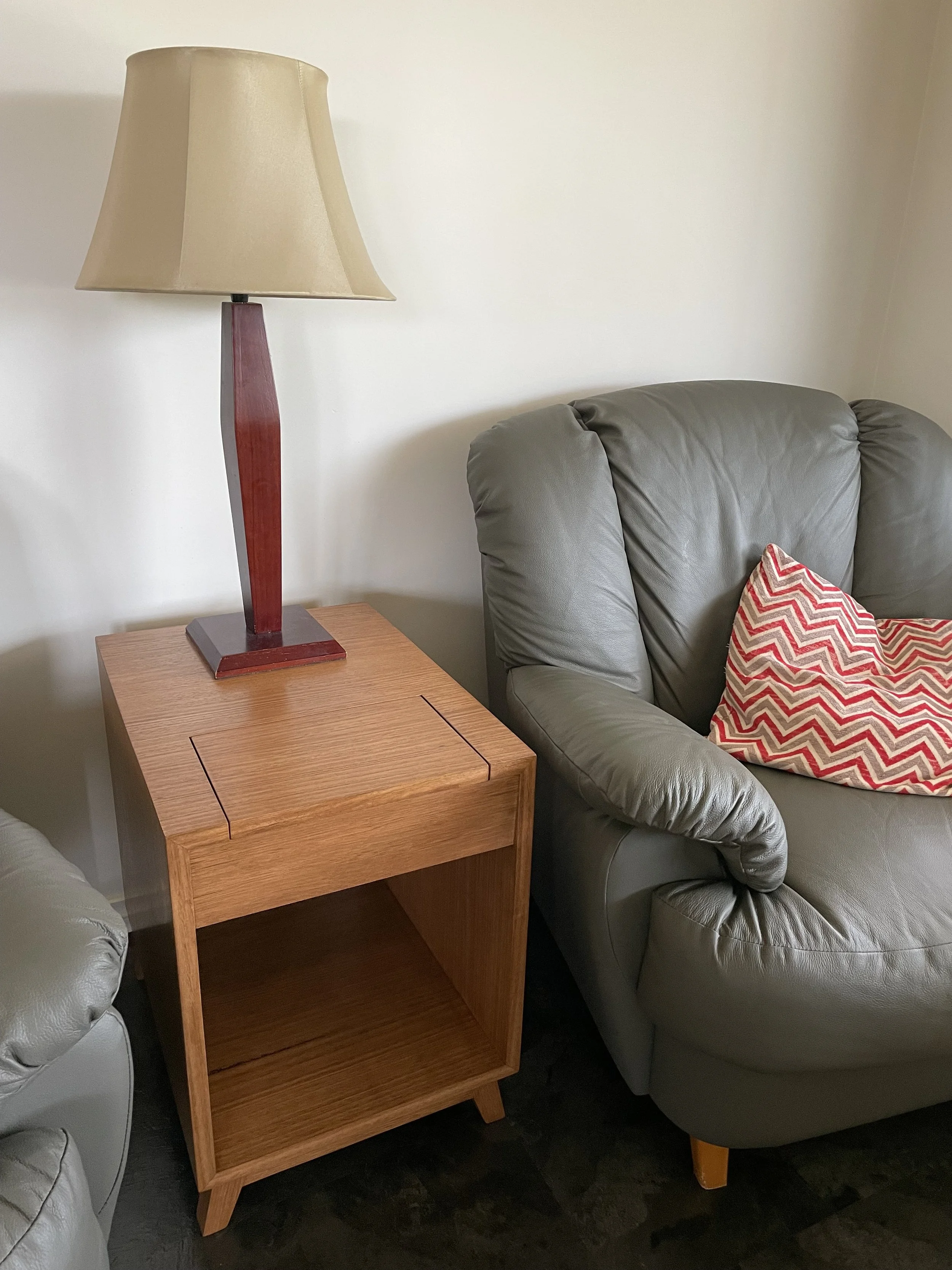 A wooden side table with a lamp on top, sitting beside a gray leather armchair with a red and white chevron pillow.