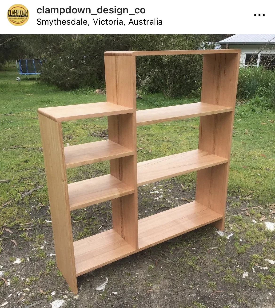 A wooden bookshelf with multiple shelves, designed with two vertical sections, placed outdoors on grass with trees and a house in the background.