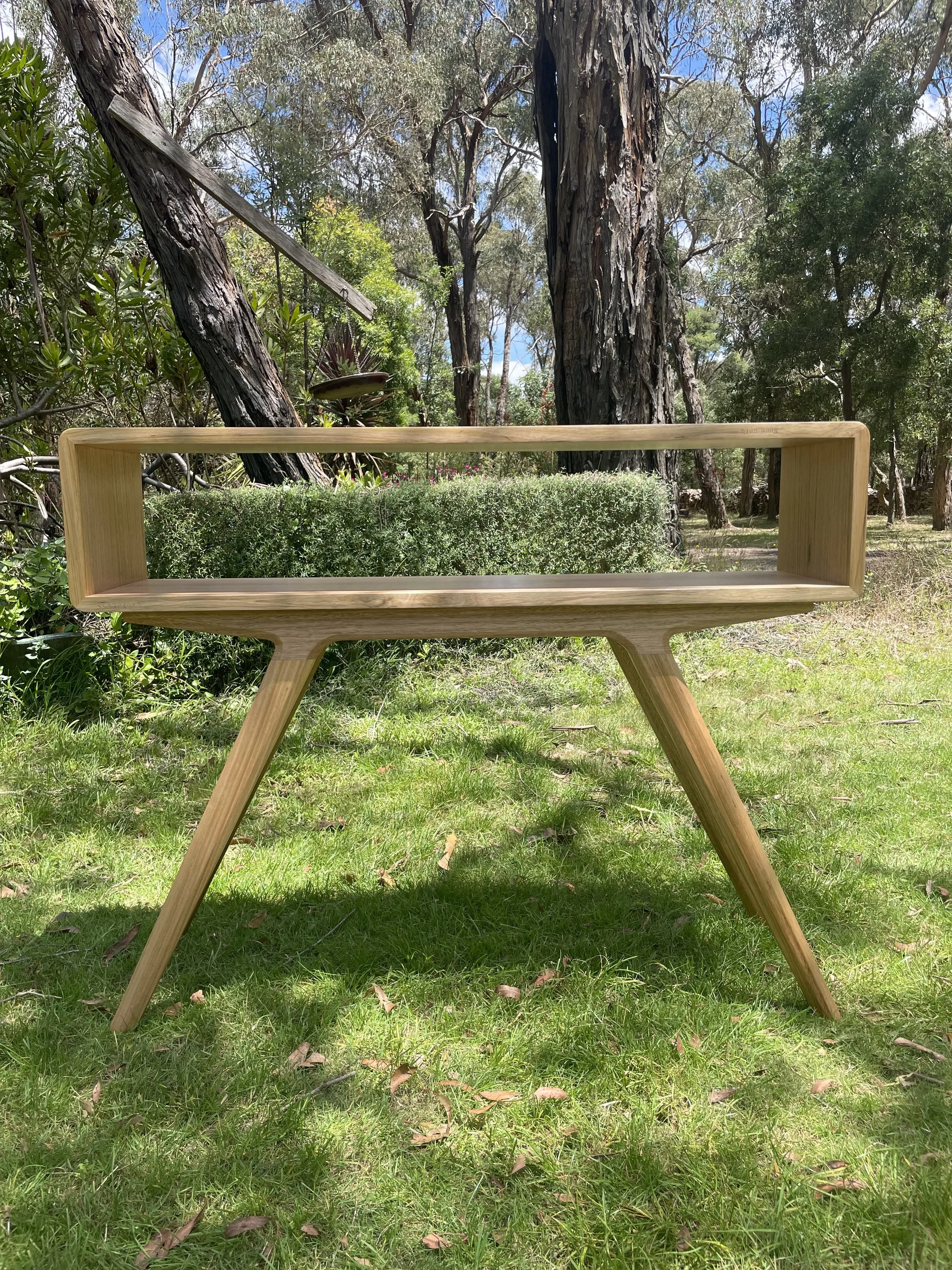 A wooden table with a rectangular open frame design, placed outdoors on grass with trees and bushes in the background.