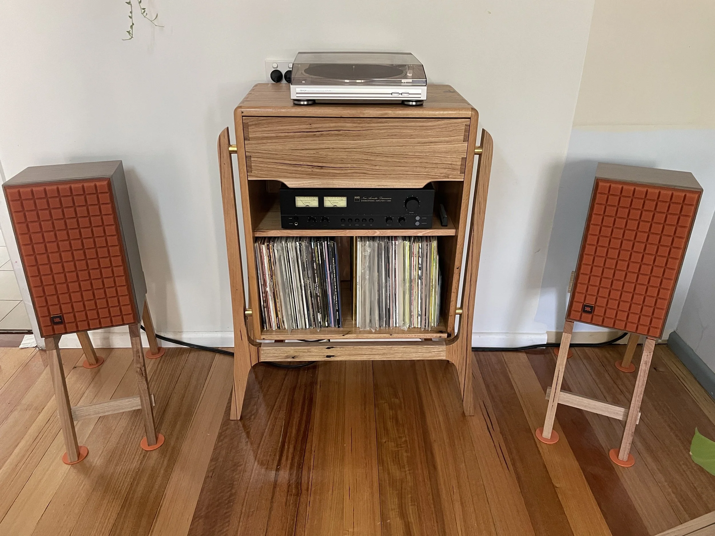 A wooden shelf with stereo equipment, vinyl records, and a turntable on top, flanked by two vintage orange and gray speakers on wooden stands on a hardwood floor.