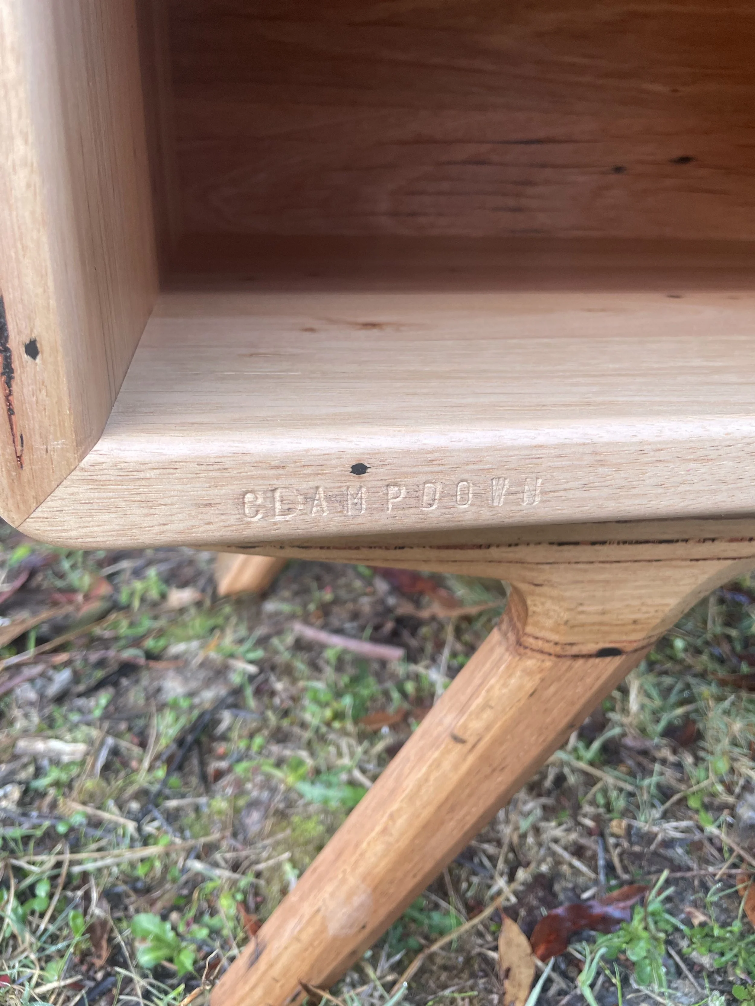 Close-up of a wooden furniture piece with an engraved stamp reading 'CEDAR PINE' on the bottom edge, outdoors on grass and leaves.