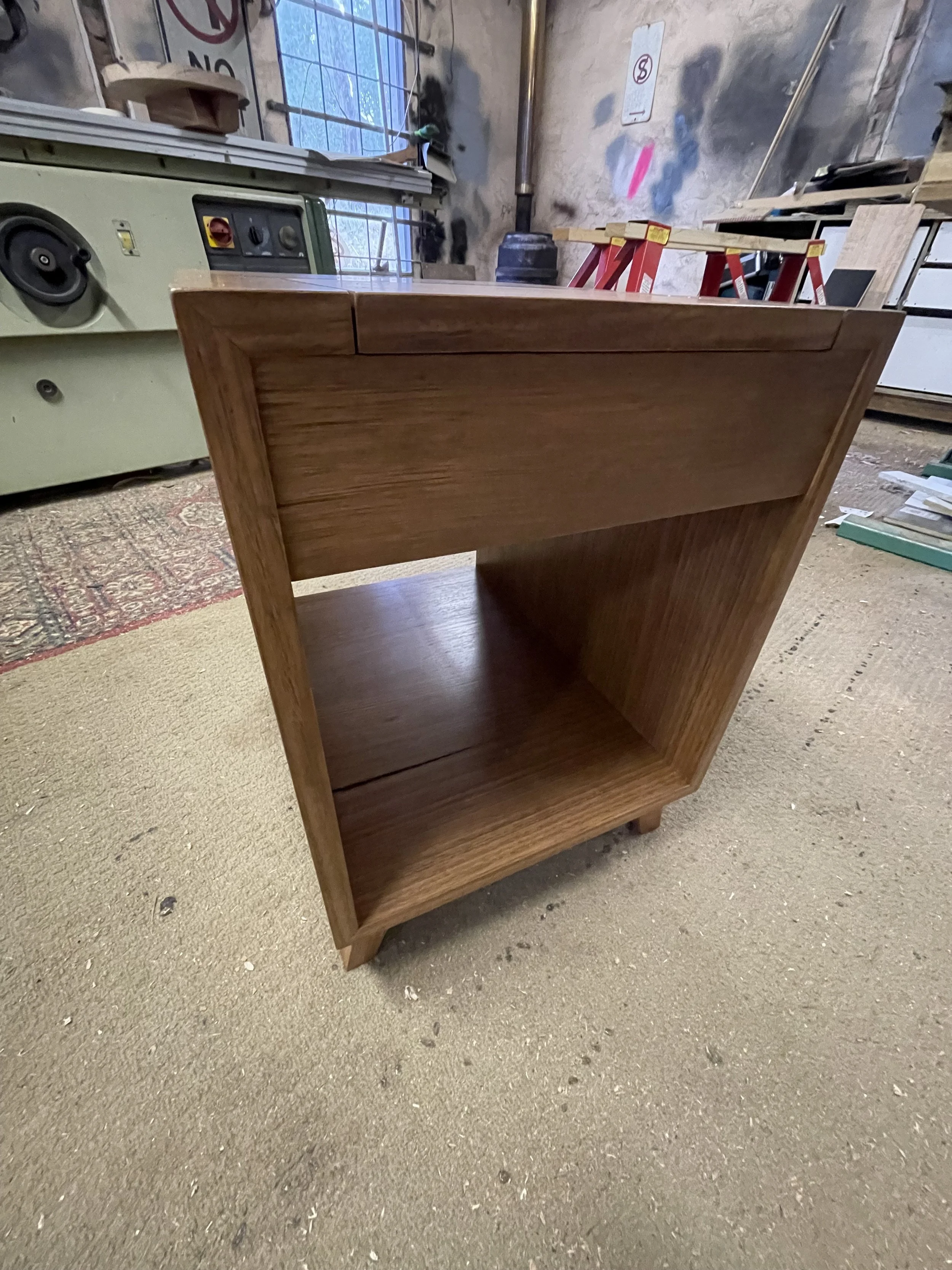 A wooden nightstand or small bookshelf in a workshop or woodshop, with tools and equipment visible in the background.