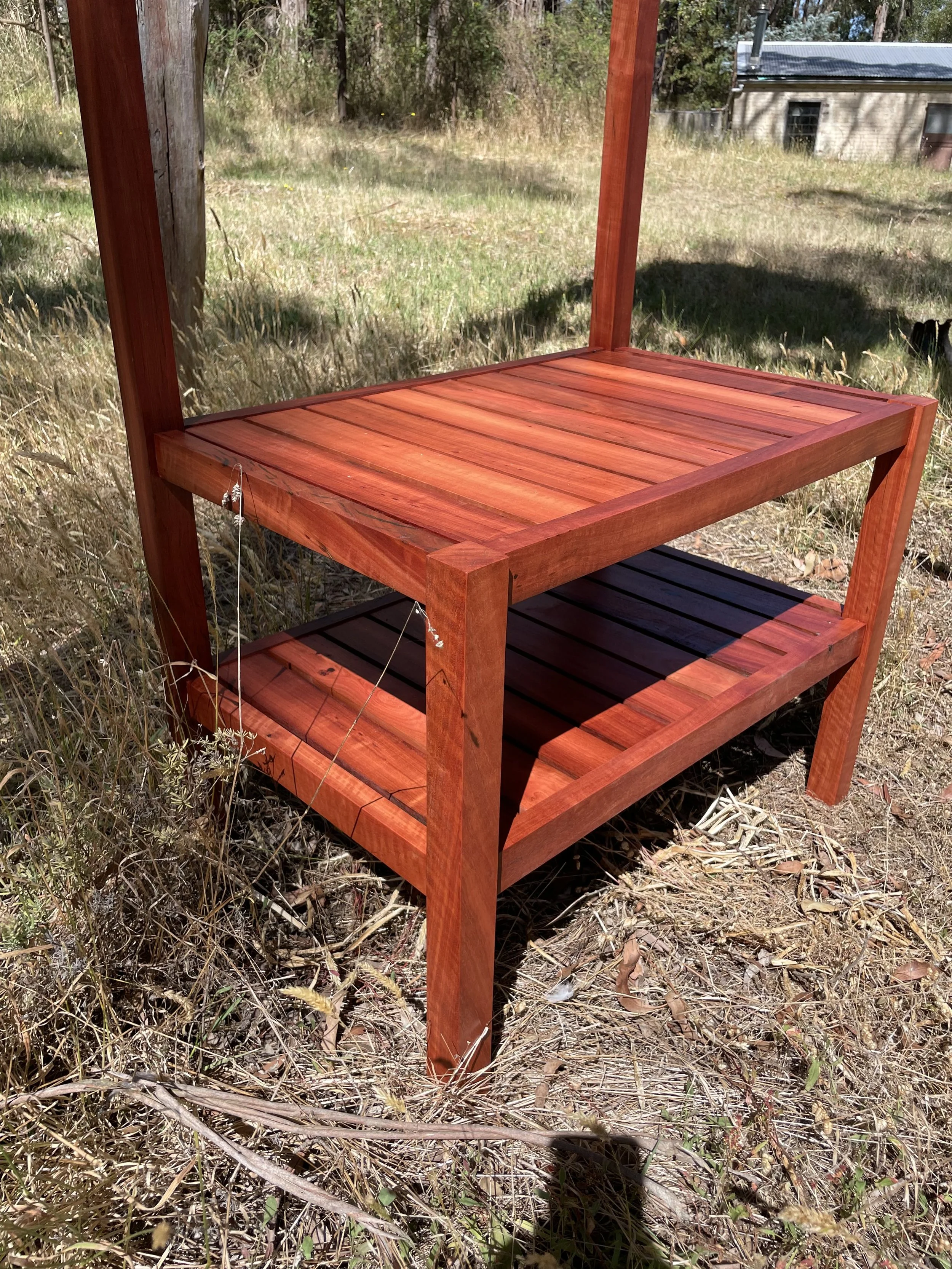 A wooden outdoor shelving unit with two shelves, situated in a grassy outdoor area with trees and a building in the background.