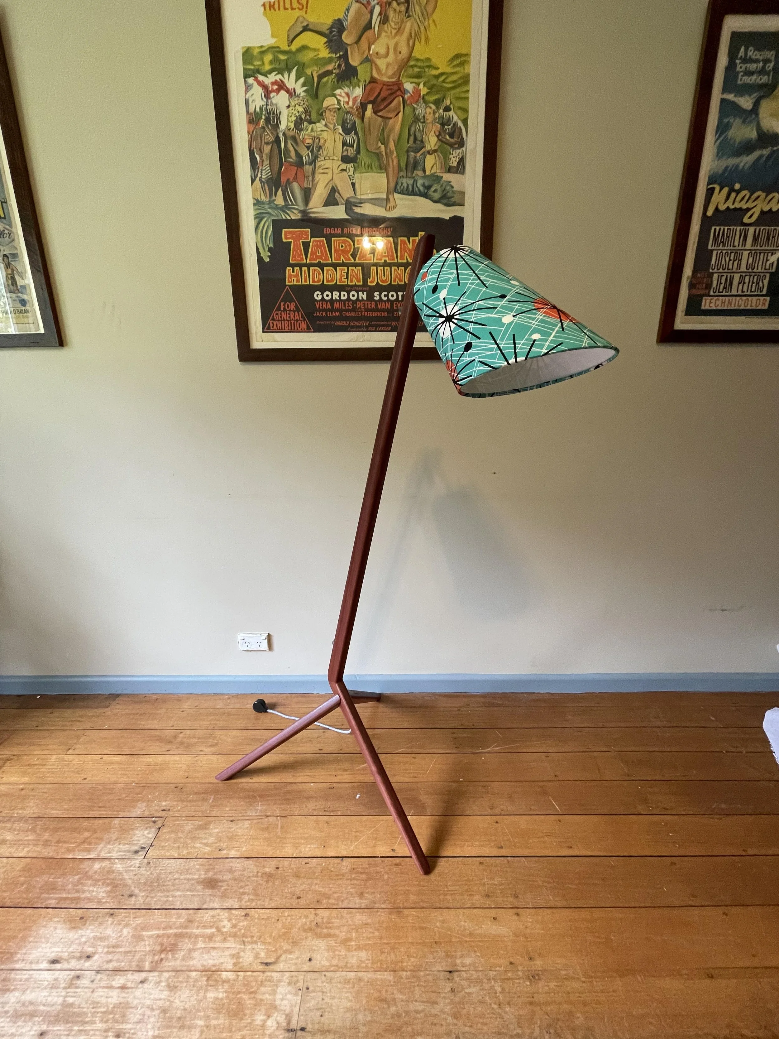A modern floor lamp with a bent wooden pole and a colorful patterned lampshade standing on a wooden floor against a beige wall decorated with framed vintage posters.