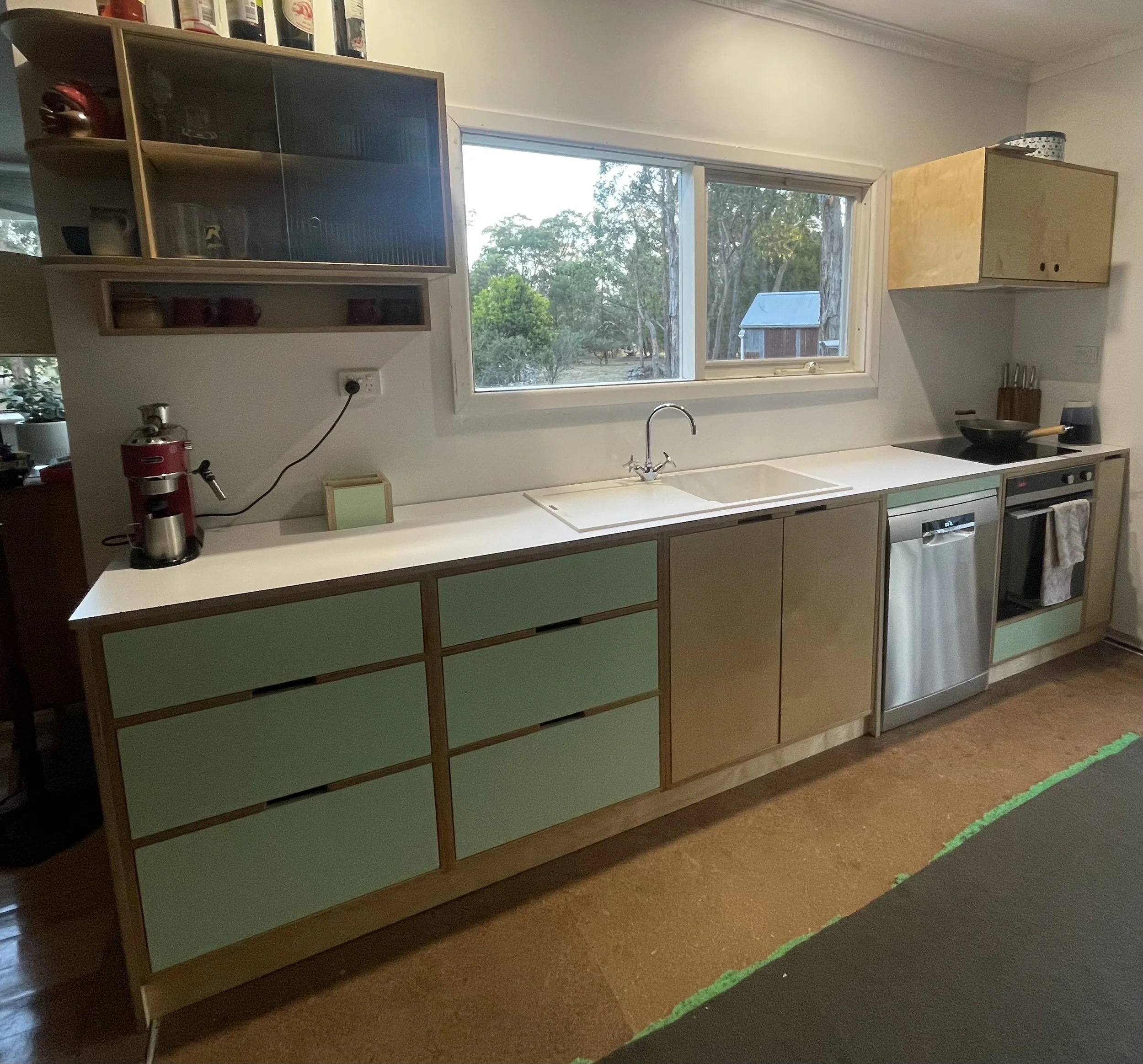 Kitchen with a white countertop, green and wooden cabinets, a window overlooking trees, a coffee machine on the counter, and a stove with a pan.