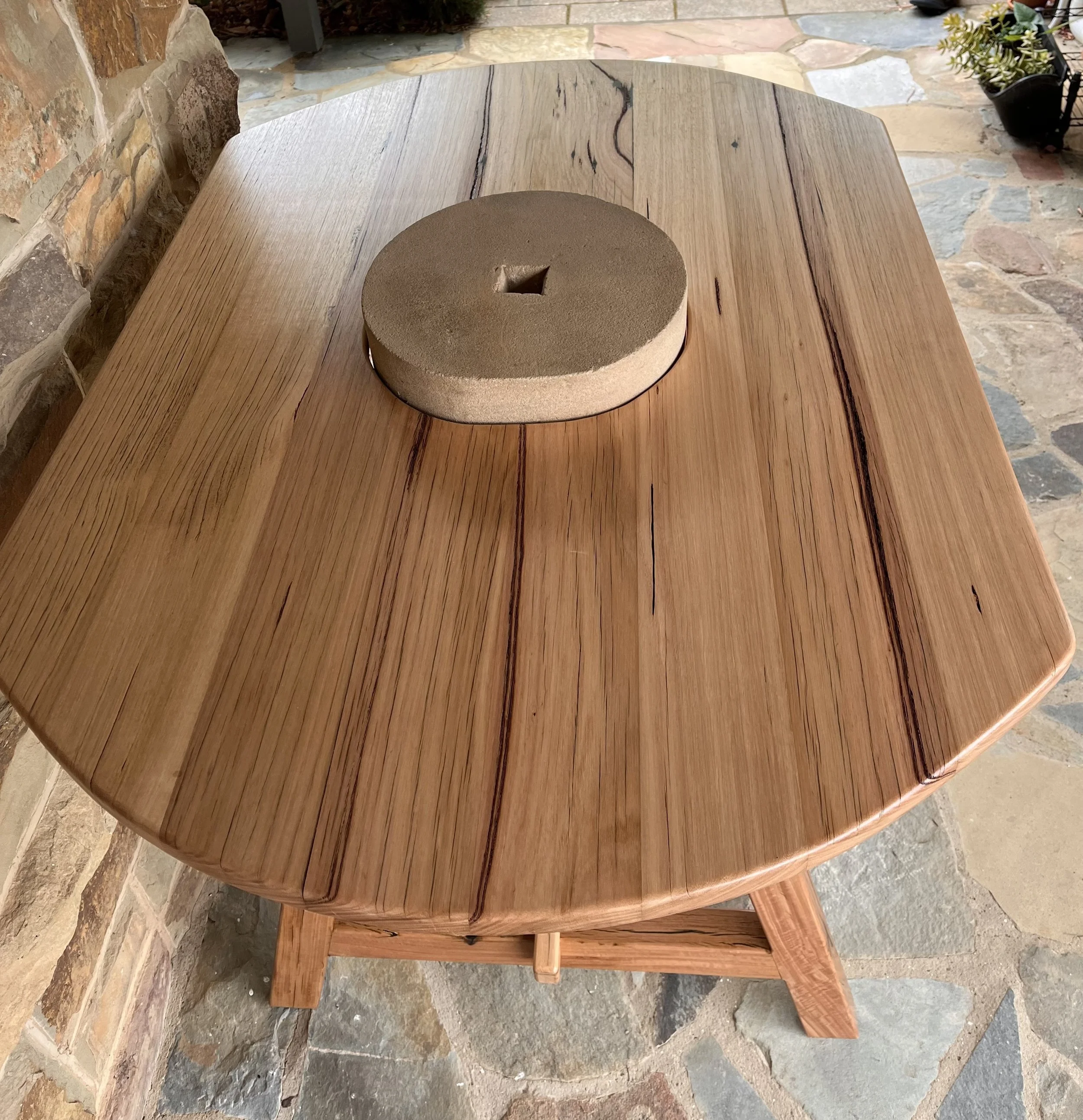 A round wooden table with a stone or ceramic object placed on top of it, situated outdoors on a stone-paved patio near a brick wall.