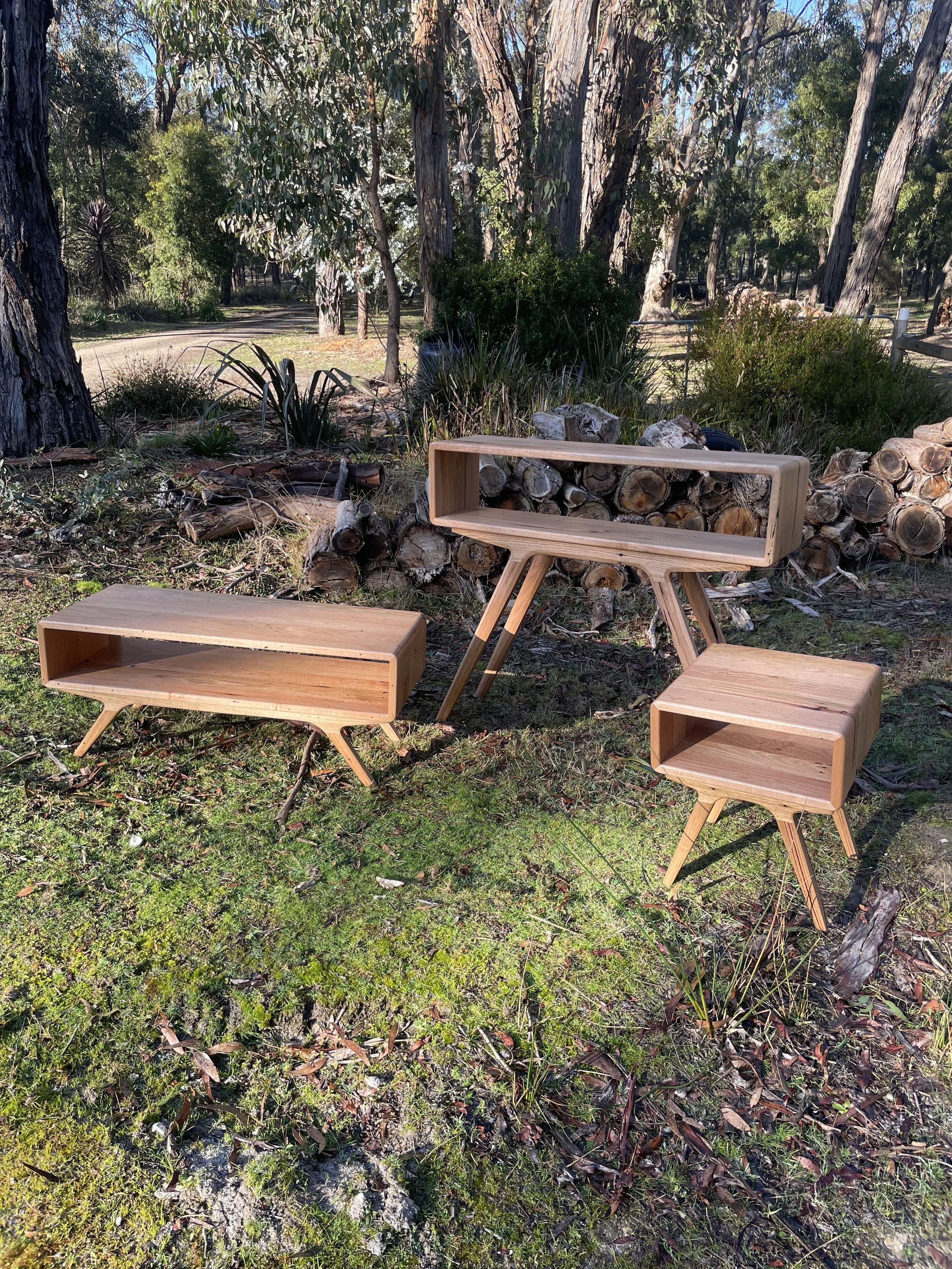 Three wooden tables with tapered legs are set outdoors on grass in a wooded area. One table is low and rectangular, another is tall and rectangular with open sides, and the third is small and square. There are logs and trees in the background.