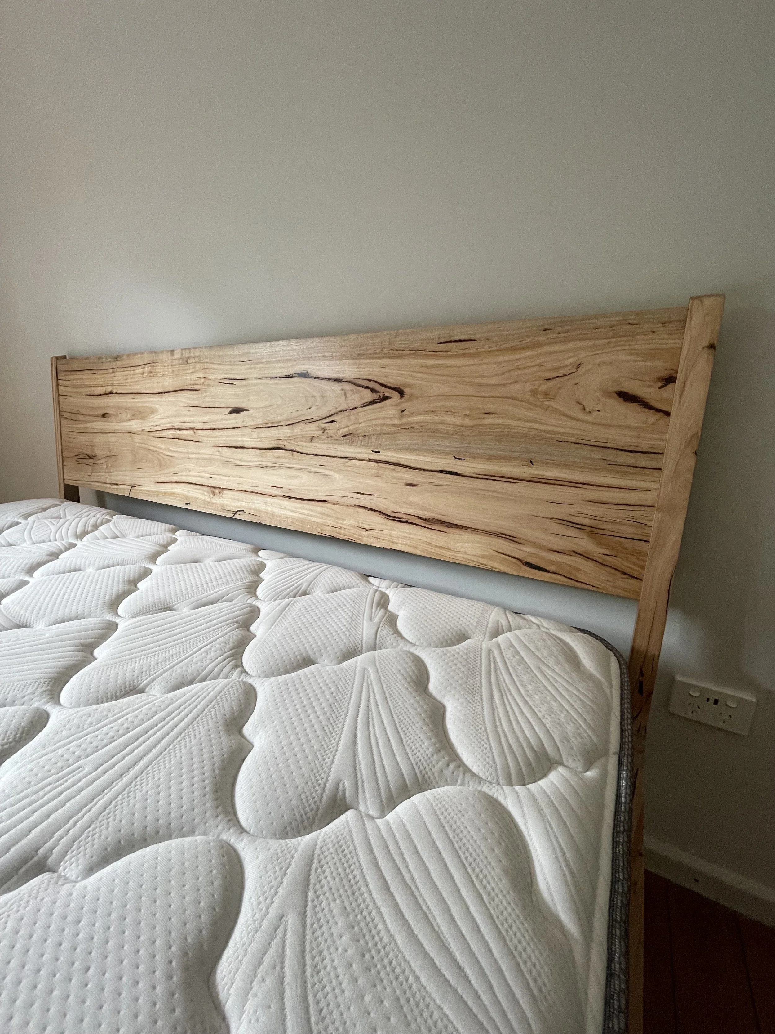 Close-up of a wooden headboard attached to a bed with a white textured mattress, next to a wall with a power outlet.