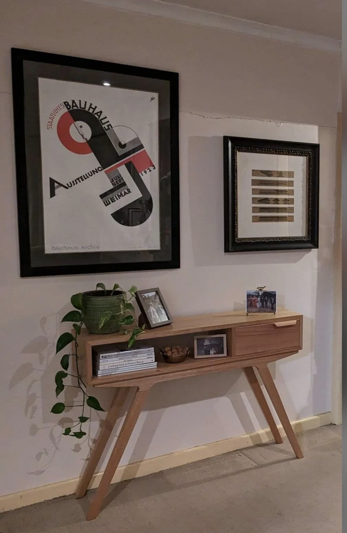 Interior wall decorated with two framed abstract posters, a wooden sideboard with pictures, a plant, and books.