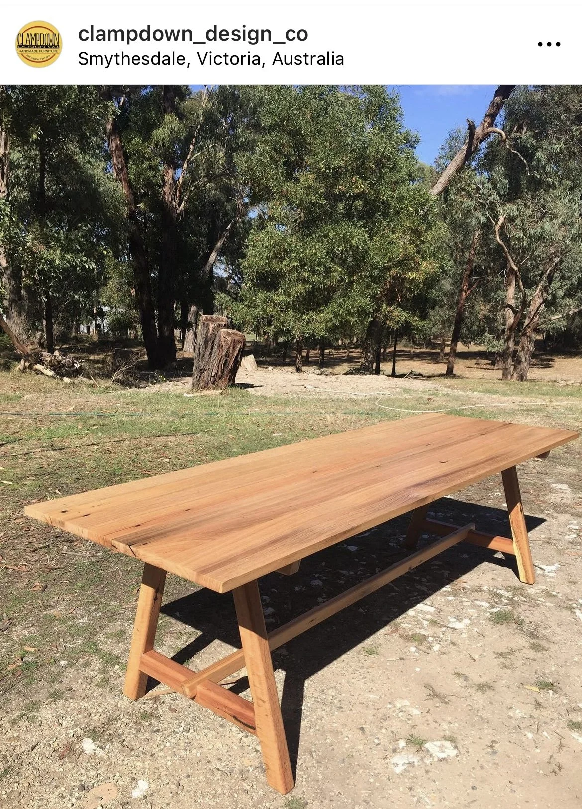 A wooden table outdoors on a dirt and grass surface, with trees and a clear blue sky in the background.