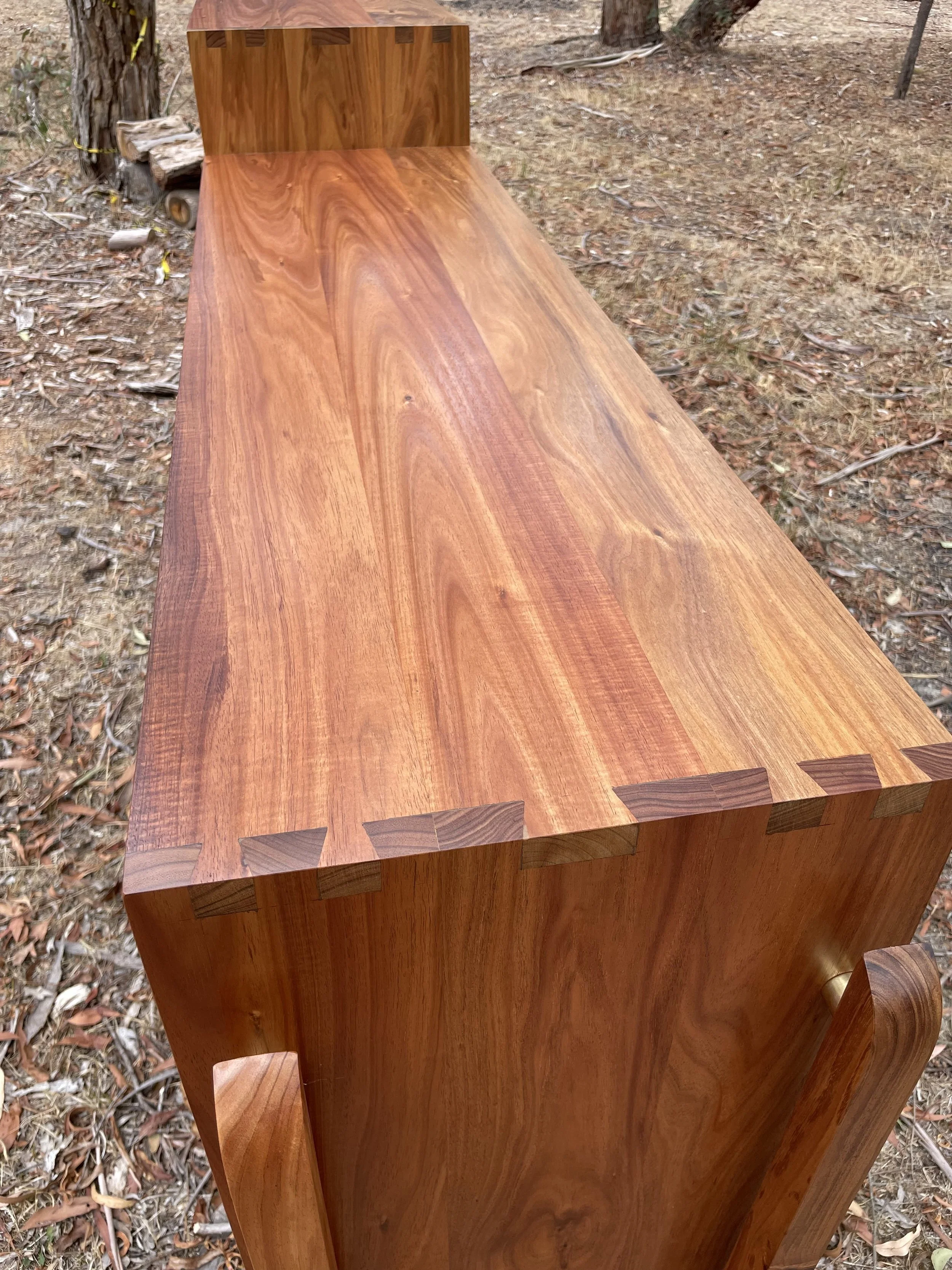 Close-up of a polished wooden bench with dovetail joints, situated outdoors on a ground with dry leaves and tree bark.