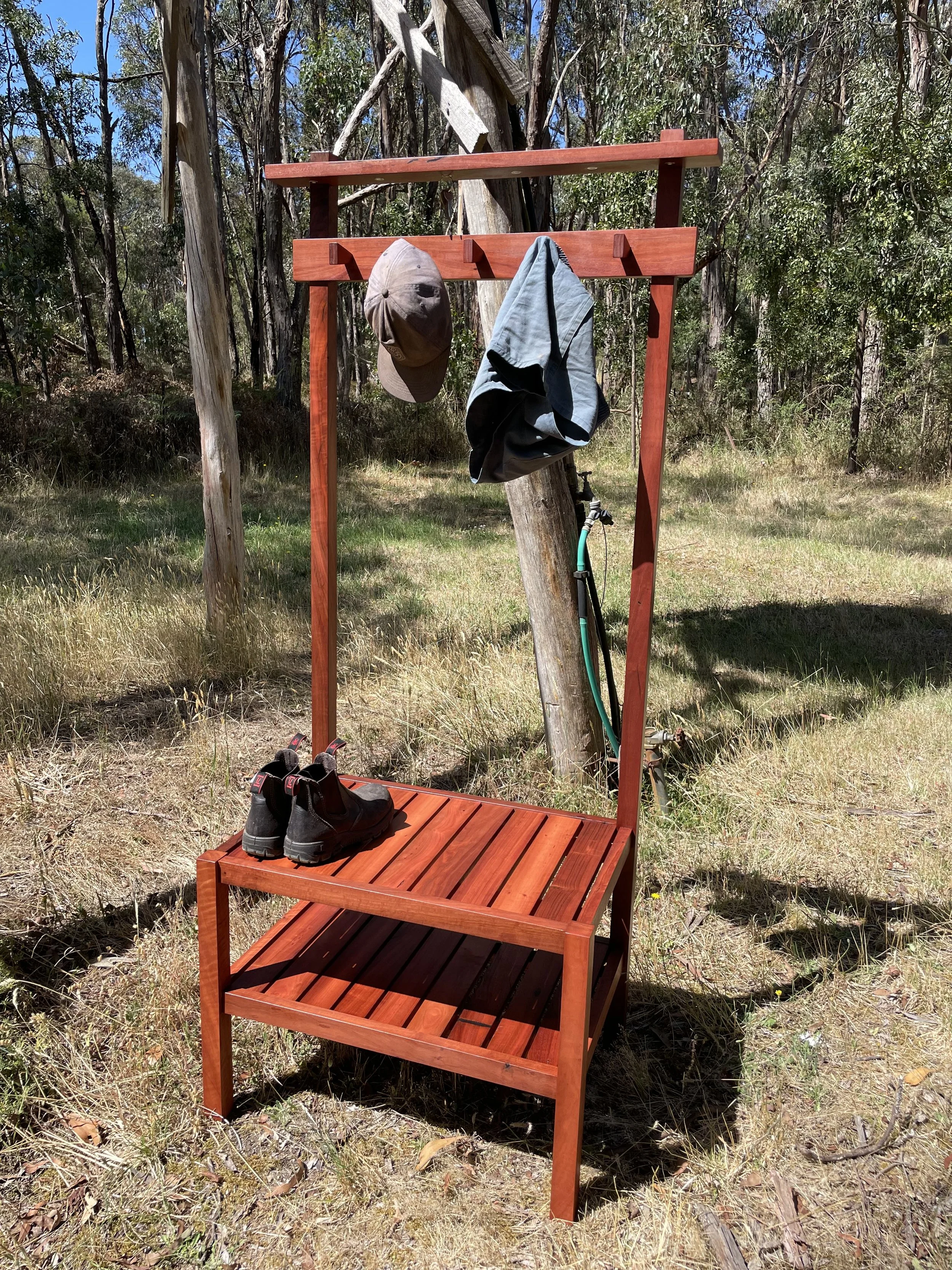 A wooden outdoor coat rack and shoe bench with a hat and a jacket hanging on it, set in a grassy area with trees in the background.