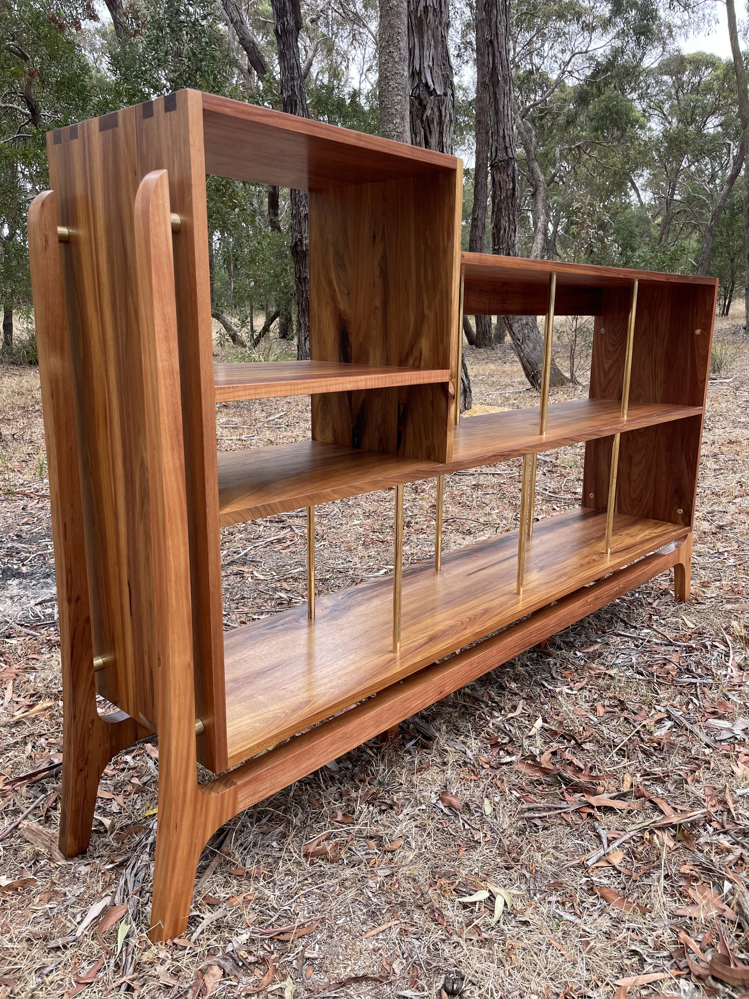 Wooden bookshelf with multiple open compartments on a dirt ground with trees in the background.