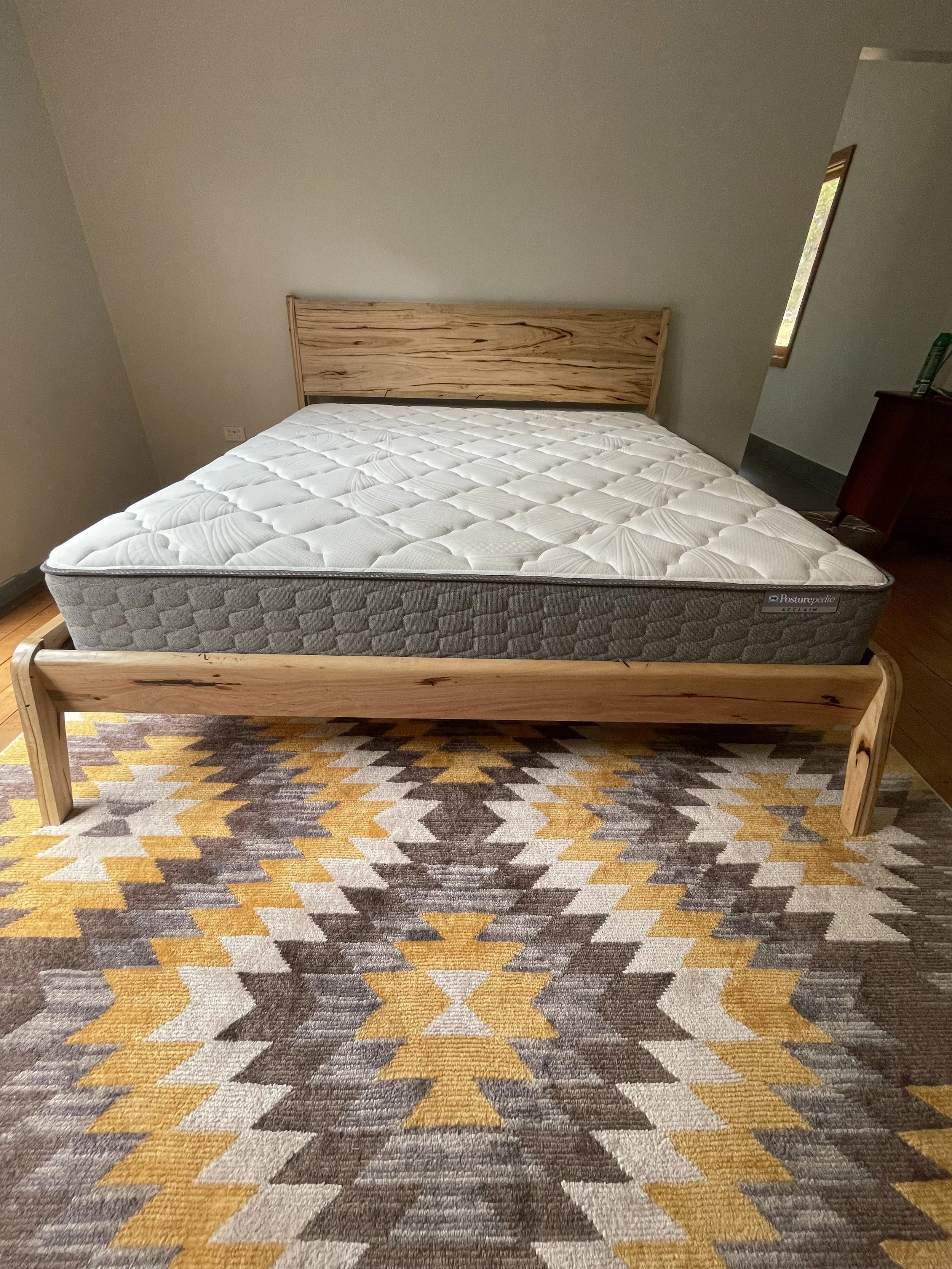 A bedroom with a wooden bed frame and a new mattress on it. The bed is positioned on a geometric patterned rug with yellow, brown, beige, and white colors. There is a window on the right side of the room.
