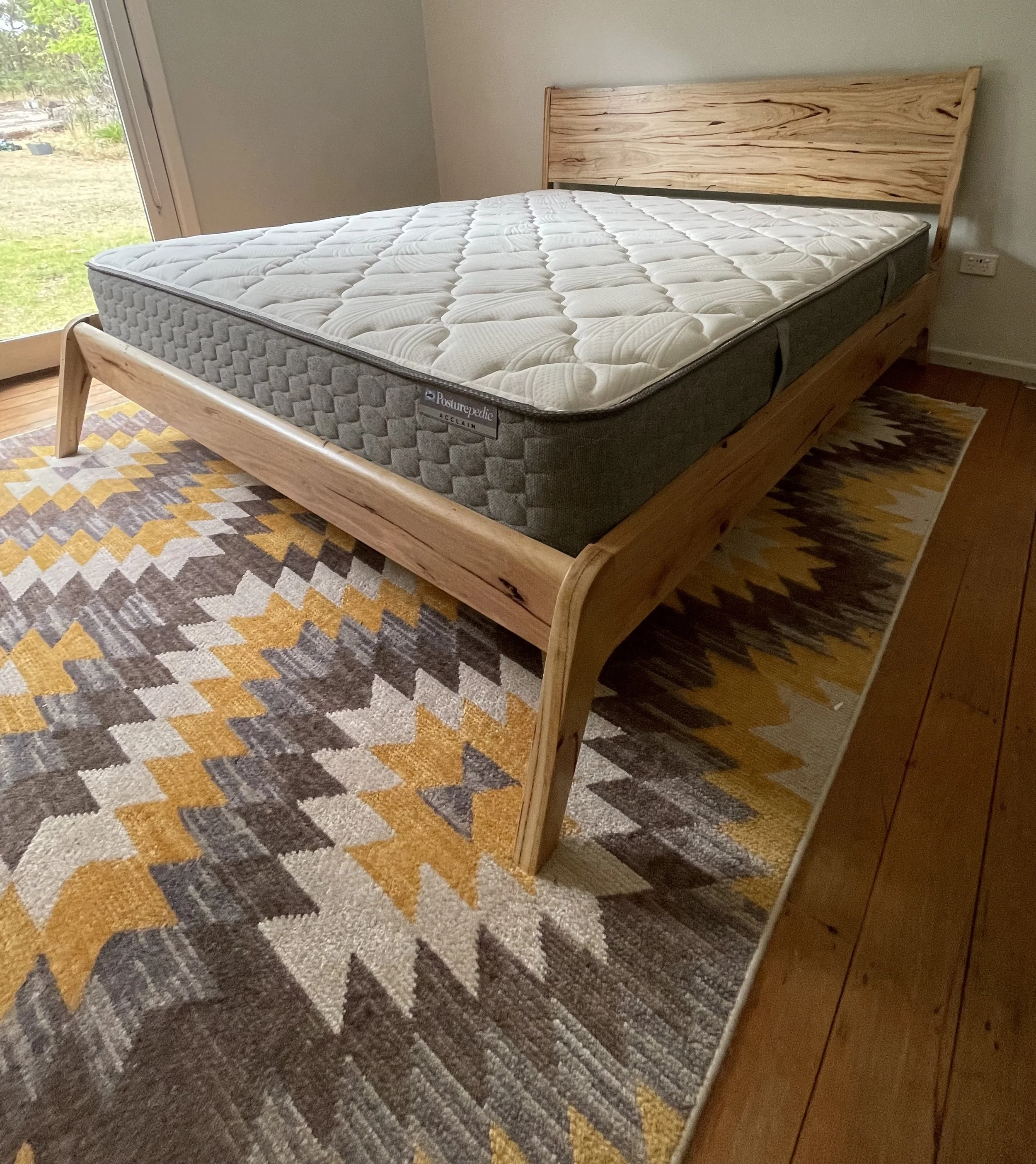 A wooden bed frame with a mattress, positioned on a colorful, patterned rug in a bedroom with a sliding glass door leading outside.