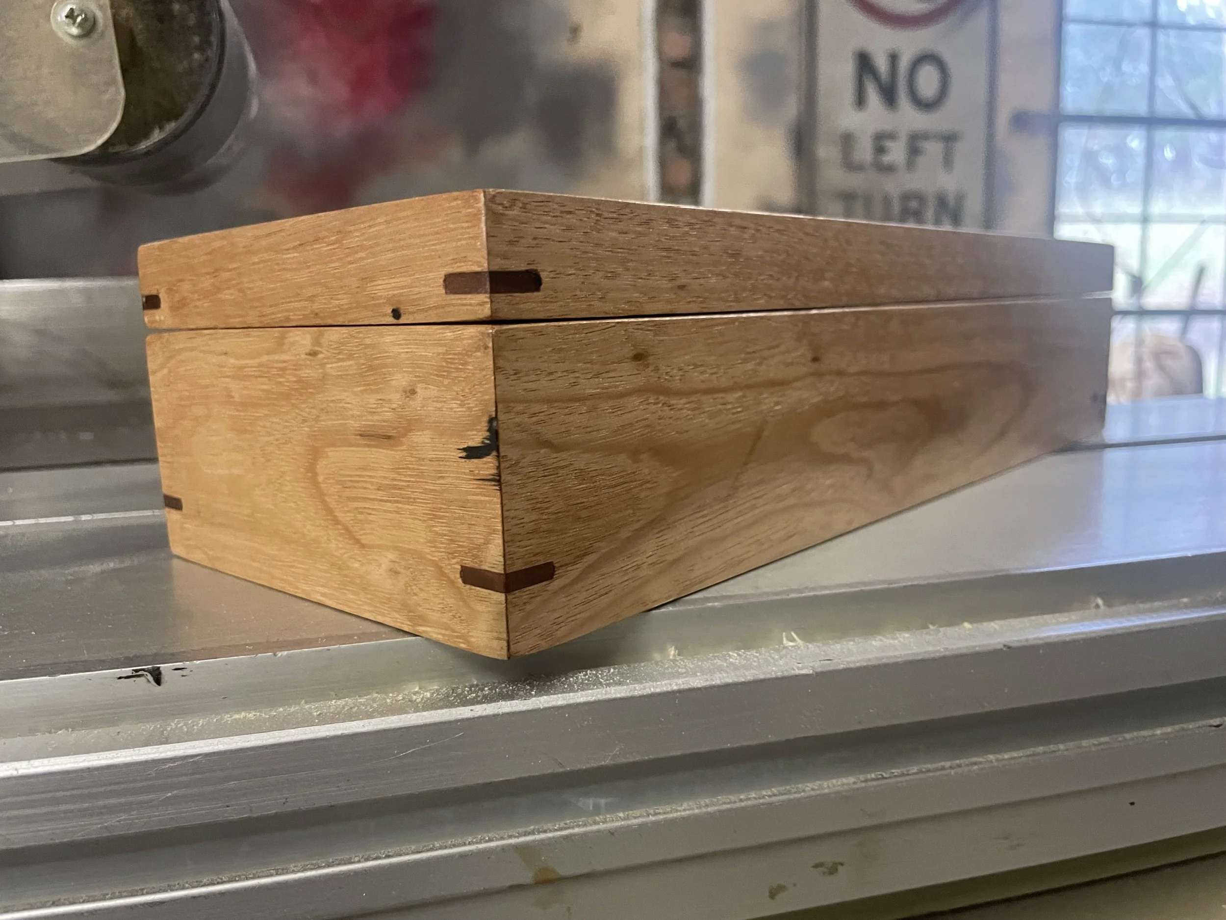 A wooden box with dovetail joints on a metal surface, with a sign in the background reading 'NO LEFT TURN'.
