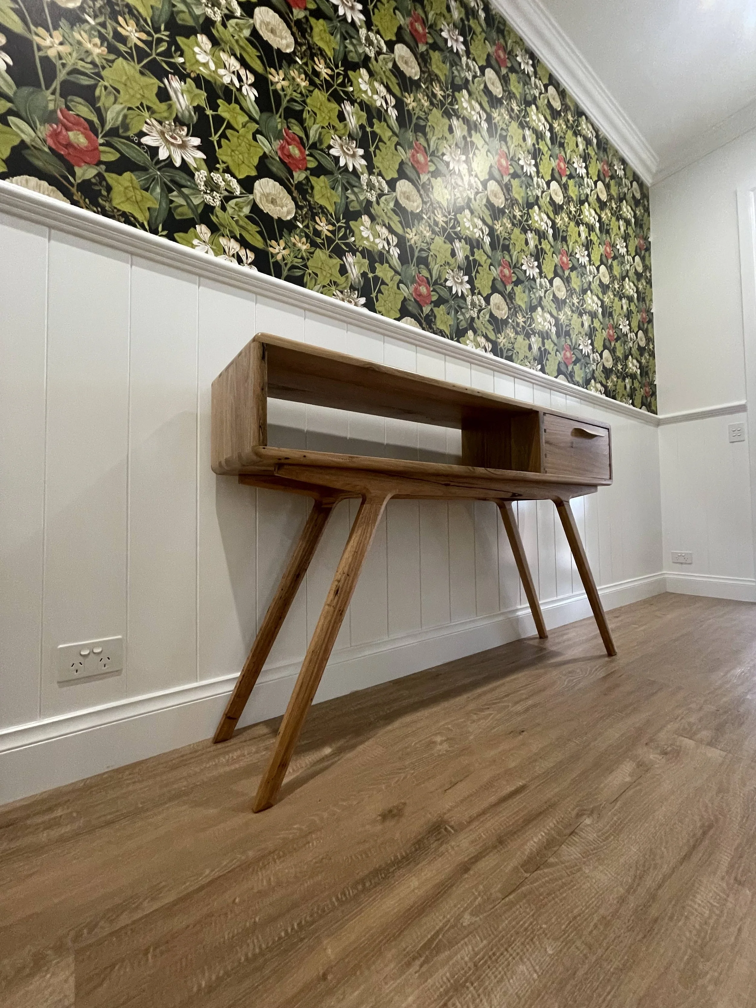 A timber hall stand with angled legs and an open shelf on the left side, placed against a wall with white wainscoting and a floral wallpaper above. The floor is made of wood, and there are electrical outlets on the wall behind the desk.