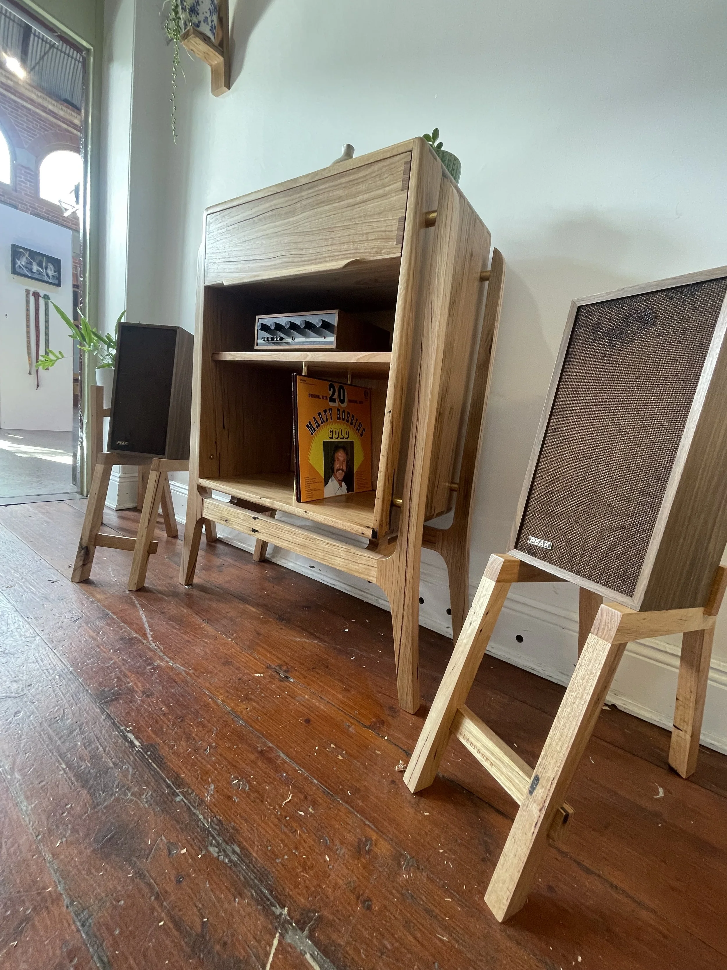 Indoor display with two wooden speakers on stands and a wooden shelf with a vinyl record and a small box, next to a window with sunlight streaming in.