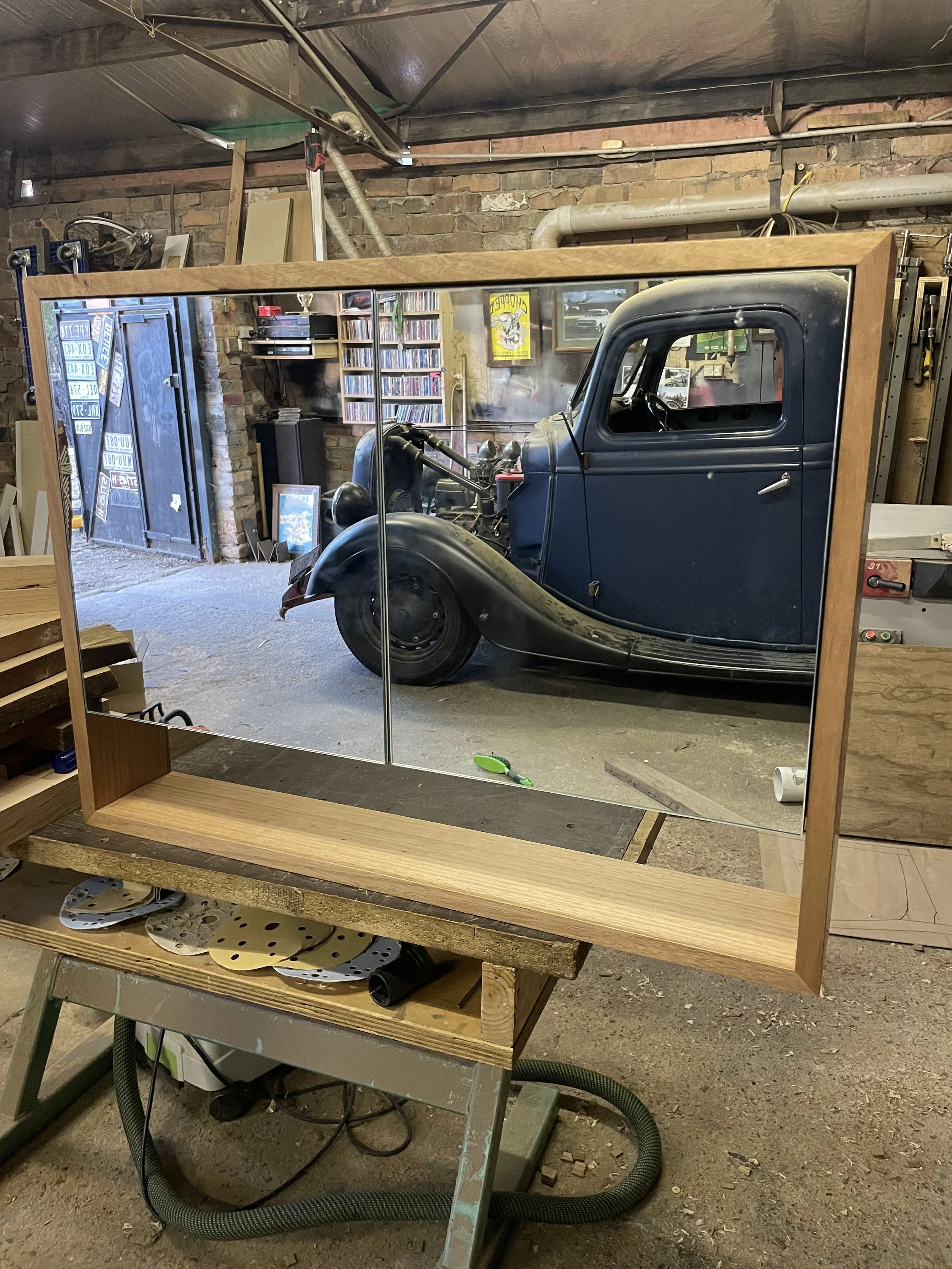 A woodworking workspace reflecting in a large mirror, showing a vintage blue car in a garage, with shelves, posters, and assorted tools and materials in the background.