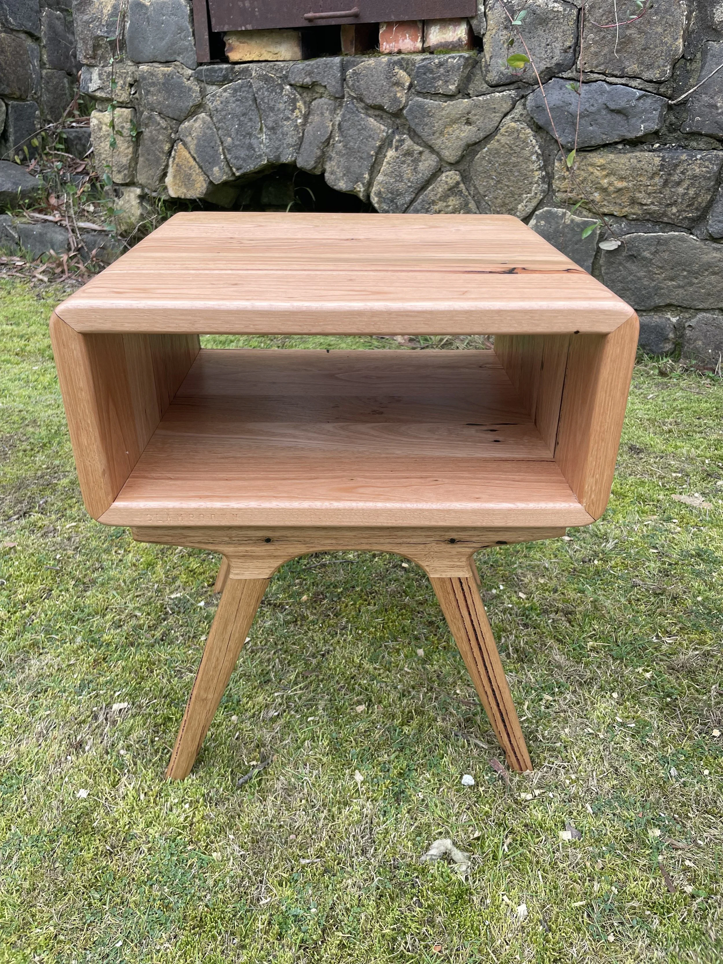 A wooden side table with slanted legs, placed on grass in front of a stone wall with a small arched opening.