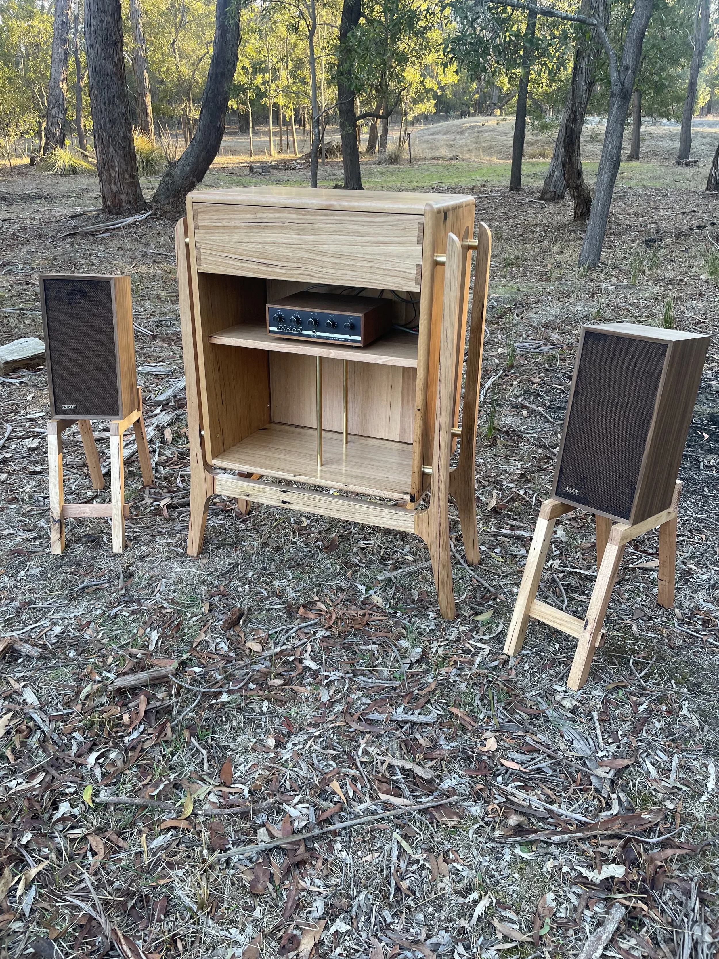 A wooden speaker cabinet with two vintage-style speakers on stands and a small amplifier inside, set outdoors in a wooded area with trees and dry leaves on the ground.