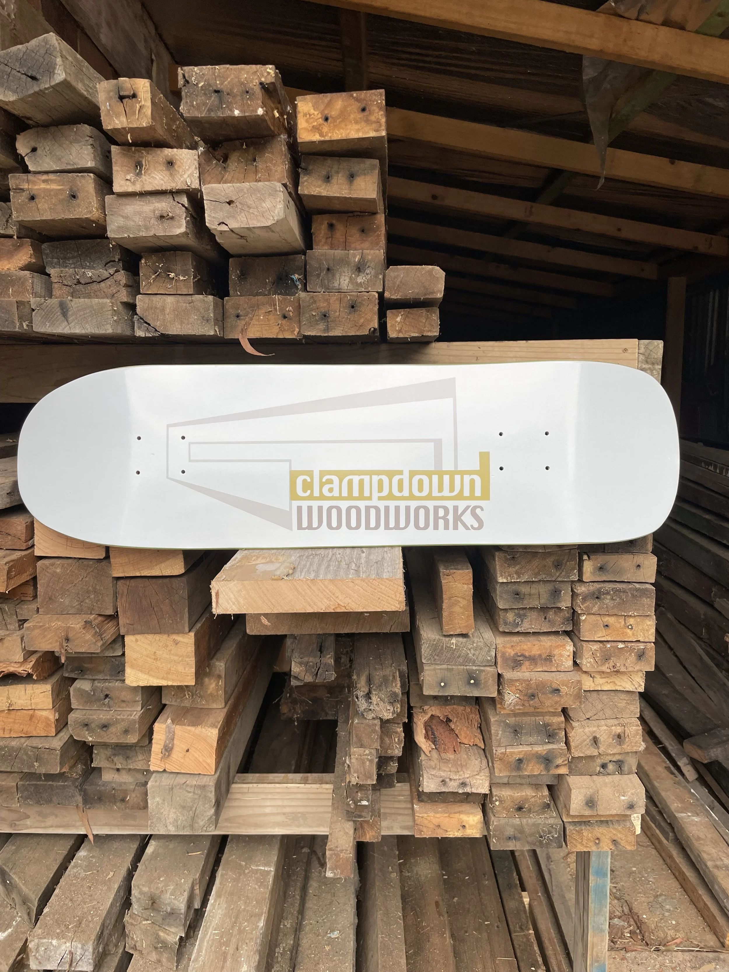 A skateboard deck with the logo and name "Clamdown Woodworks" leaning against a stack of wooden planks in a woodworking storage area.