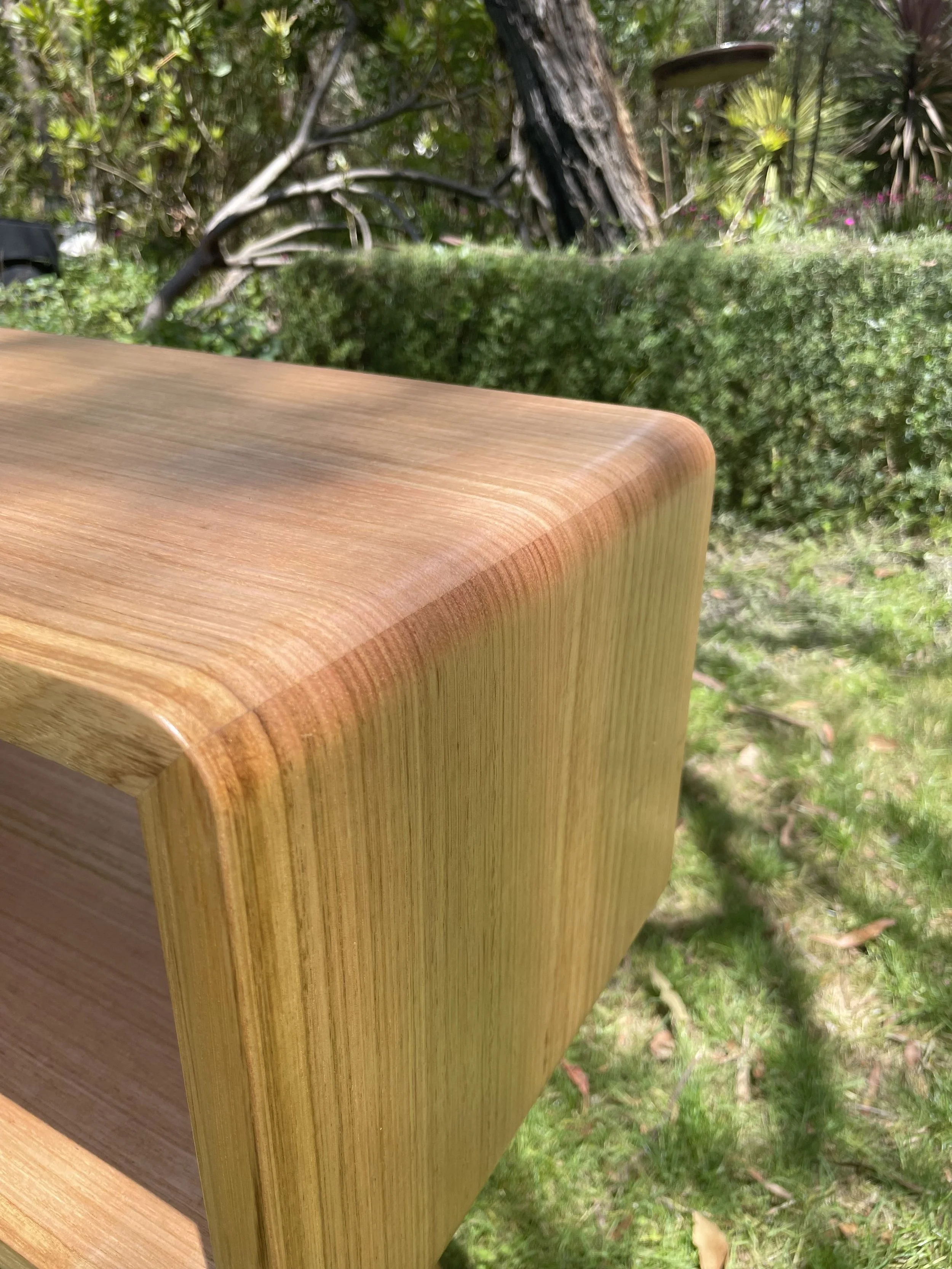 Close-up of a wooden piece of furniture, likely a table or shelf, outdoors with trees, bushes, and grass in the background.