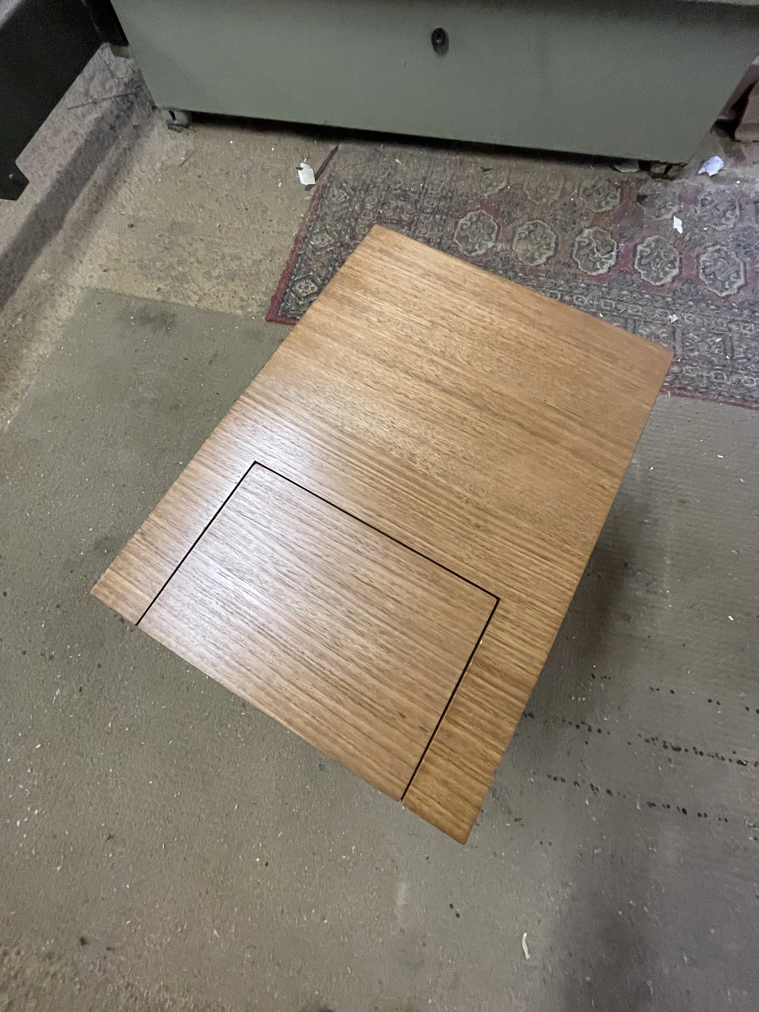 A wooden table with a compartment in the middle, placed on a concrete floor next to a decorative rug.