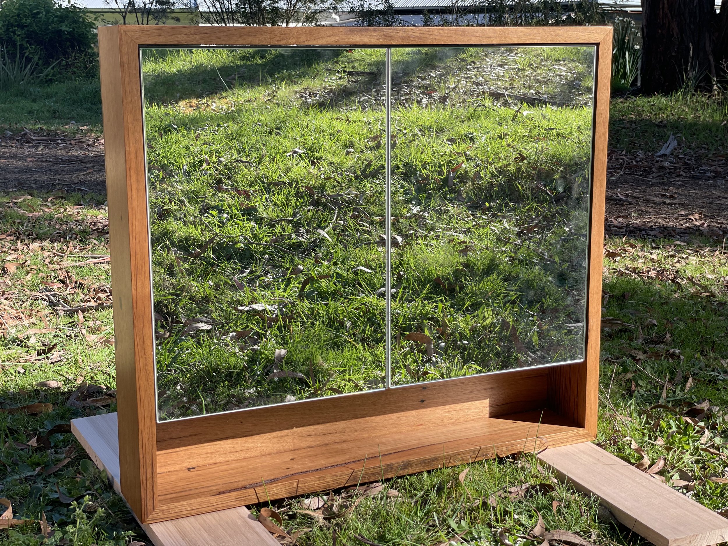 A wooden framed mirror with a small shelf at the bottom, placed outdoors on a grassy area.