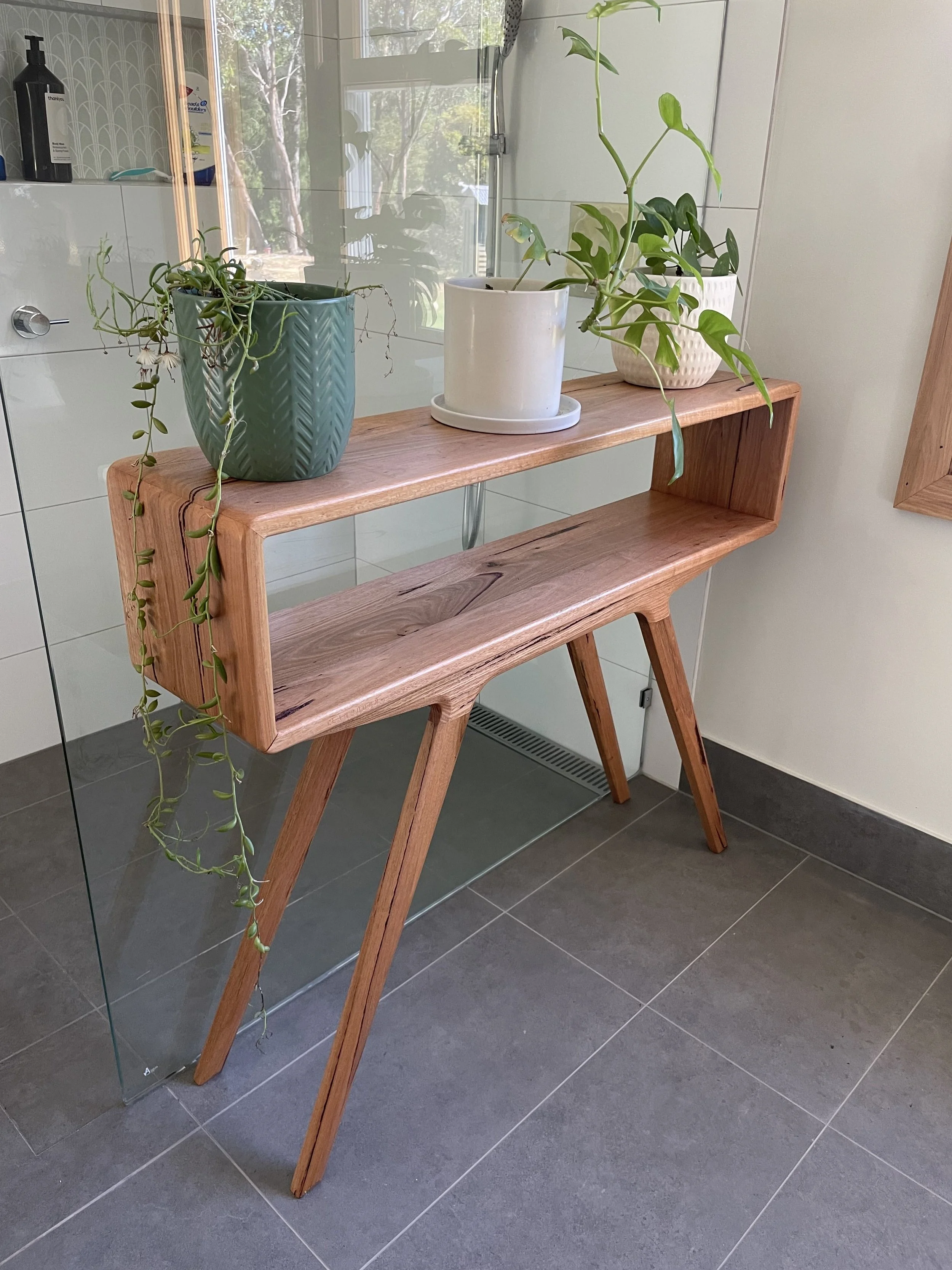 Three potted plants on a wooden shelf inside a bathroom, with a glass shower enclosure and tiled wall in the background.
