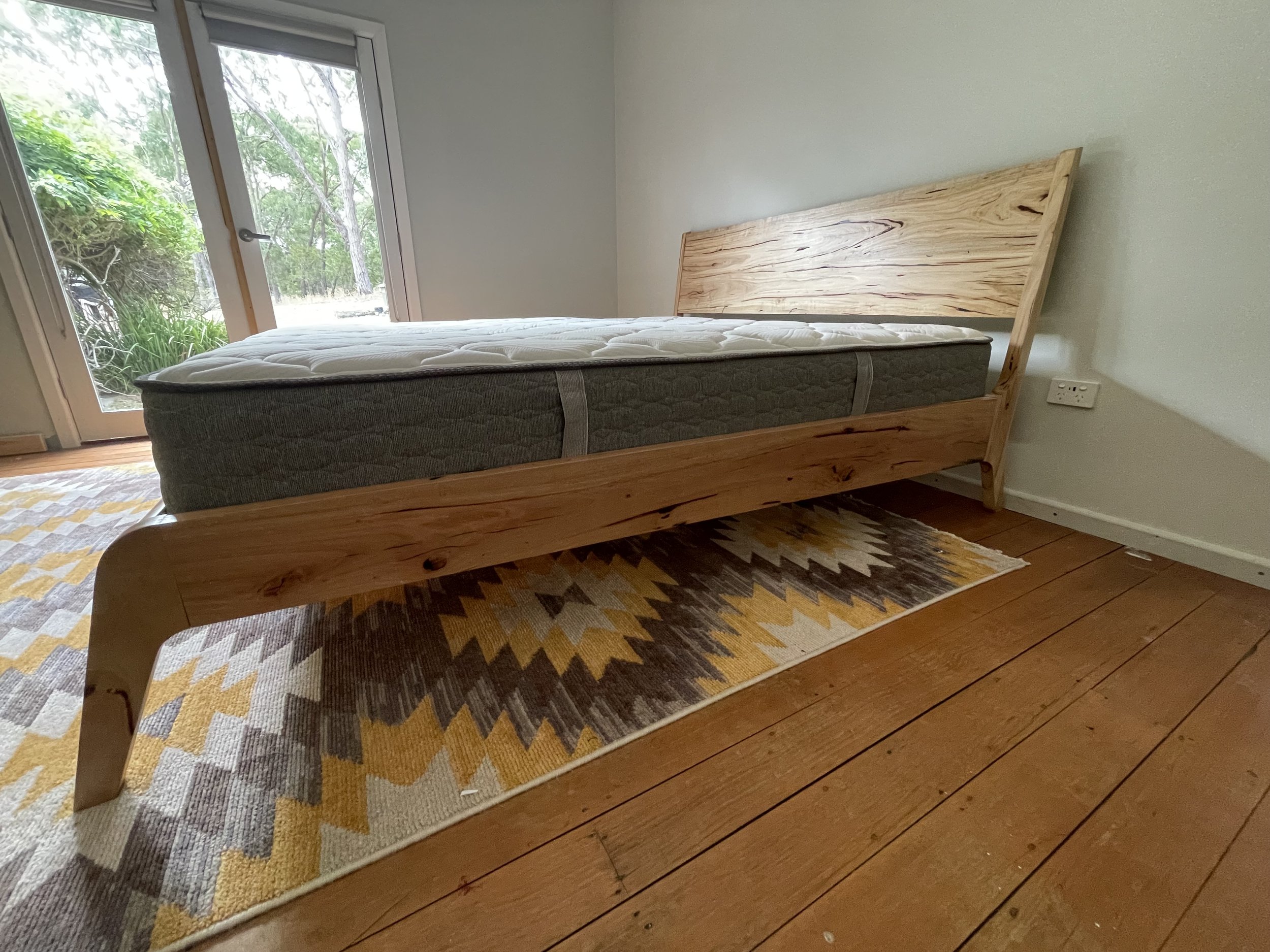 A bed with a wooden frame and a mattress in a room with wooden floors and a patterned rug. There is a glass door leading outside to greenery.