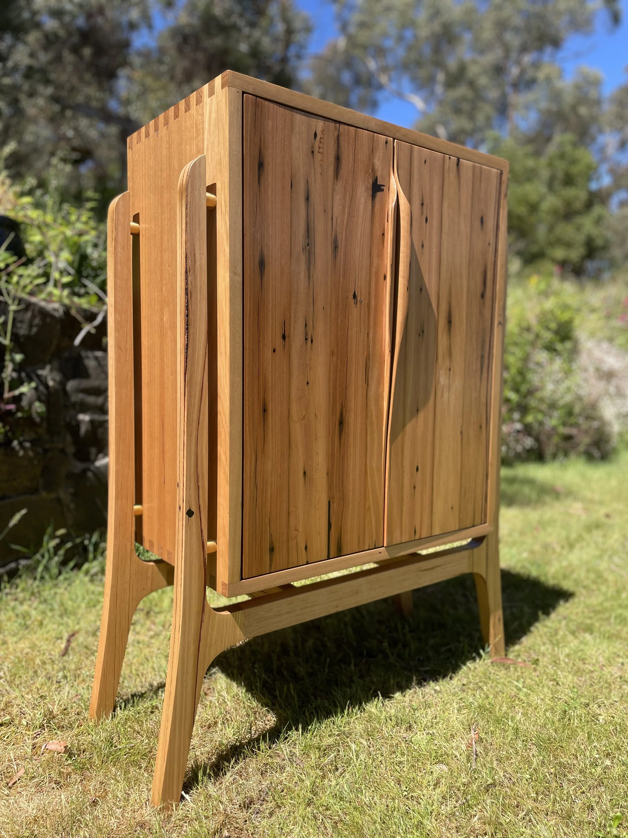 A wooden cabinet with slatted doors, standing outdoors on a grassy area with trees in the background.