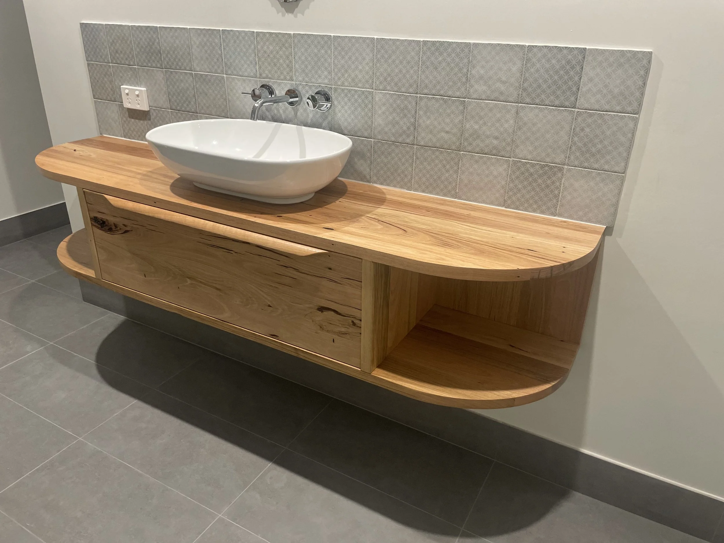 Wooden bathroom vanity with an oval vessel sink and wall-mounted faucet, set against a tiled wall with electrical outlets.