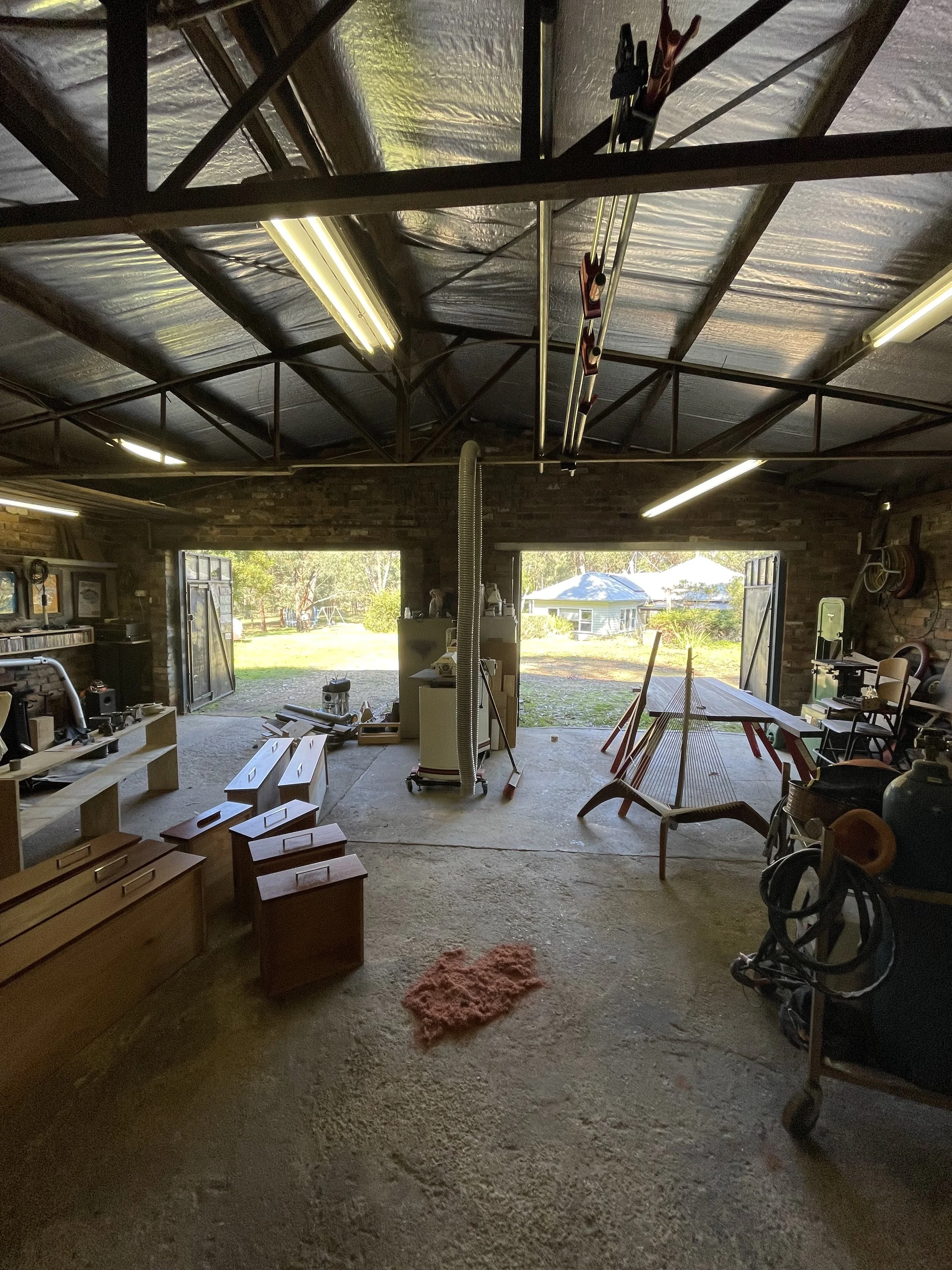 A spacious garage with open double doors showing a backyard with trees, a swing set, and a shed. The garage contains various woodworking and gardening tools, furniture pieces, and a hammock, with shelving on the right side and fluorescent lighting on