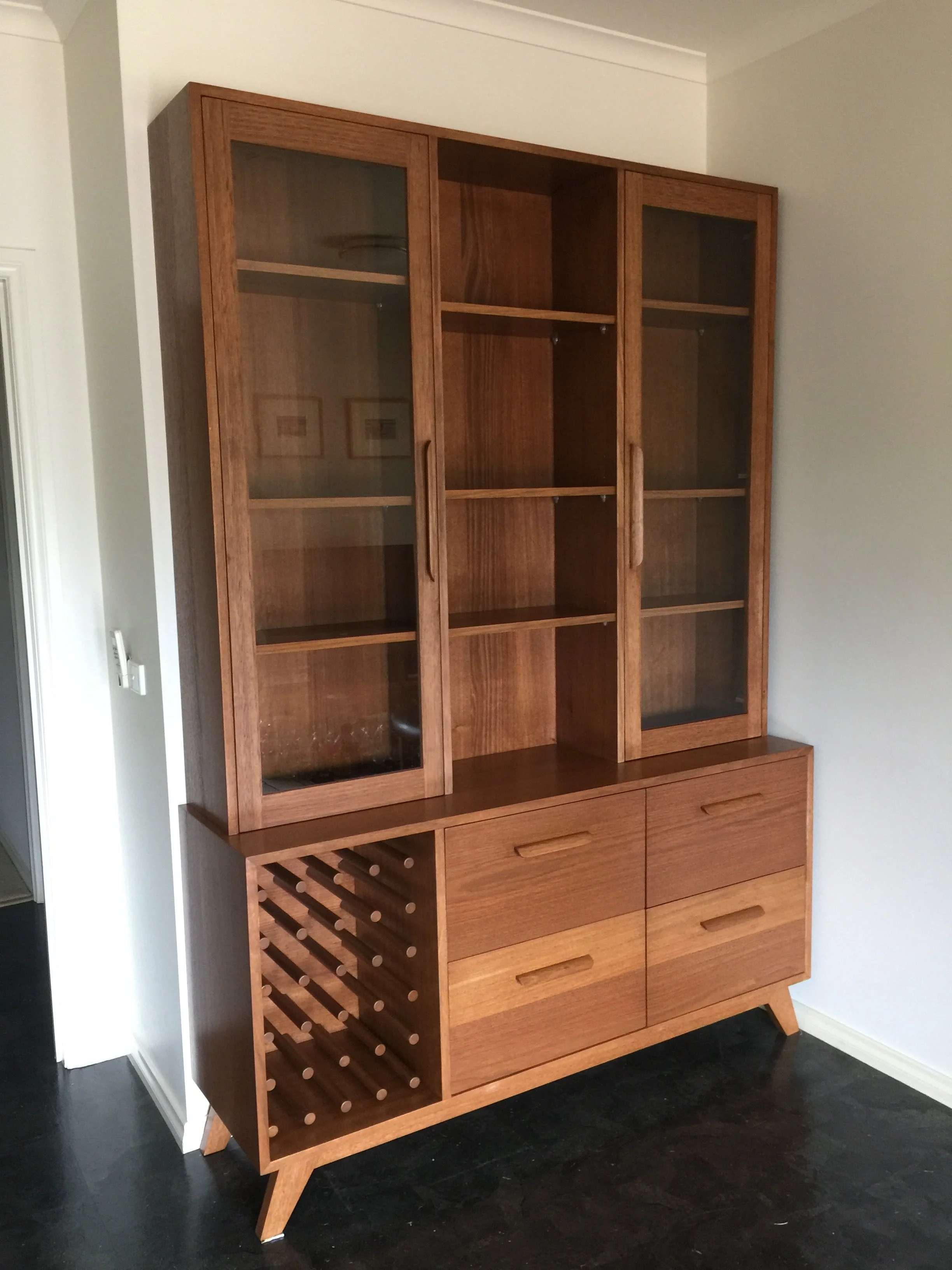 A mid-century modern wooden cabinet with glass doors on the upper section, open central shelves, and four drawers on the lower section. The left side has a built-in wine rack. The cabinet stands on four angled legs.
