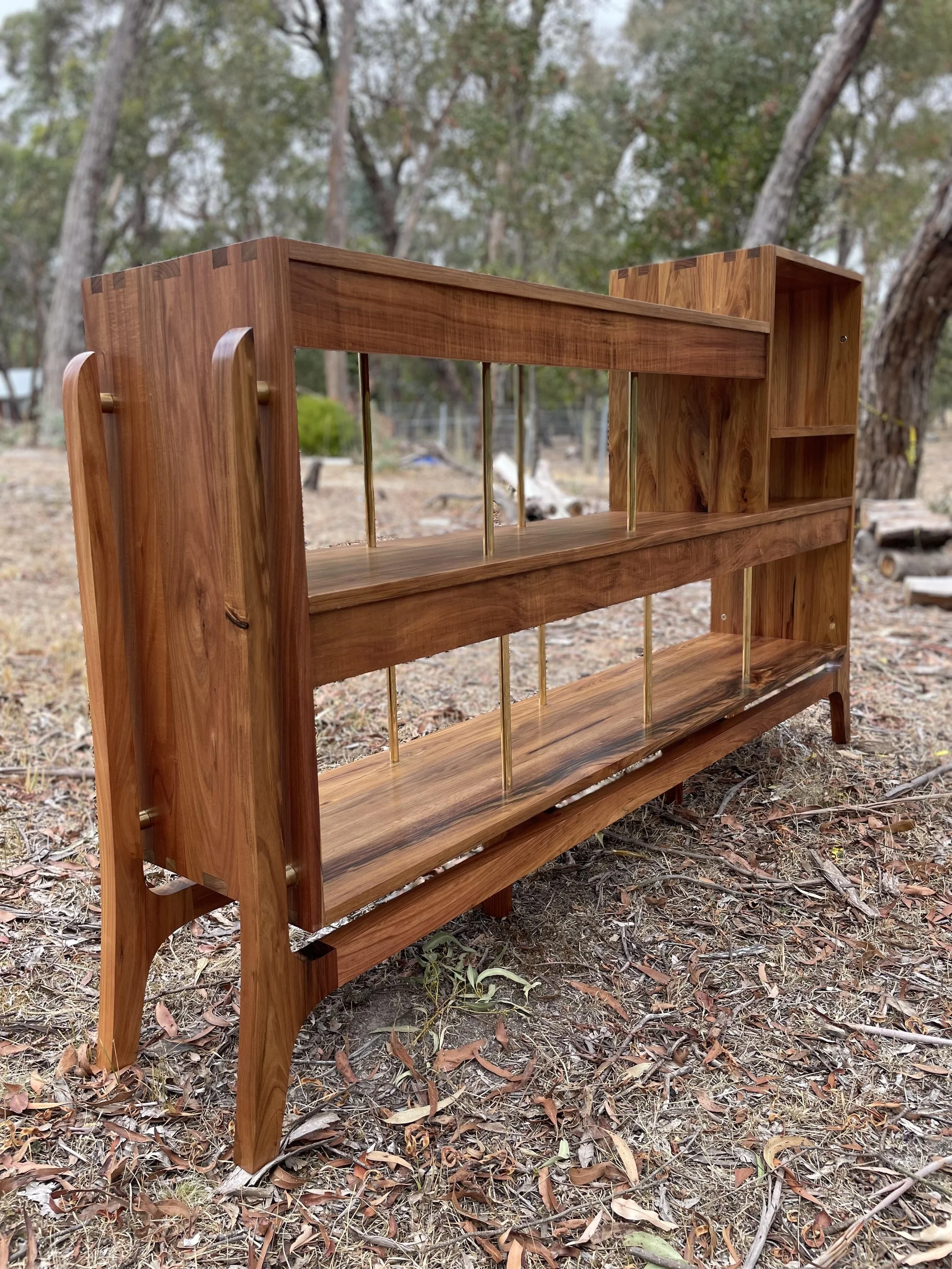 A wooden bookshelf with three tiers, gold metal rods supporting open compartments, set outdoors on a dirt ground with trees in the background.