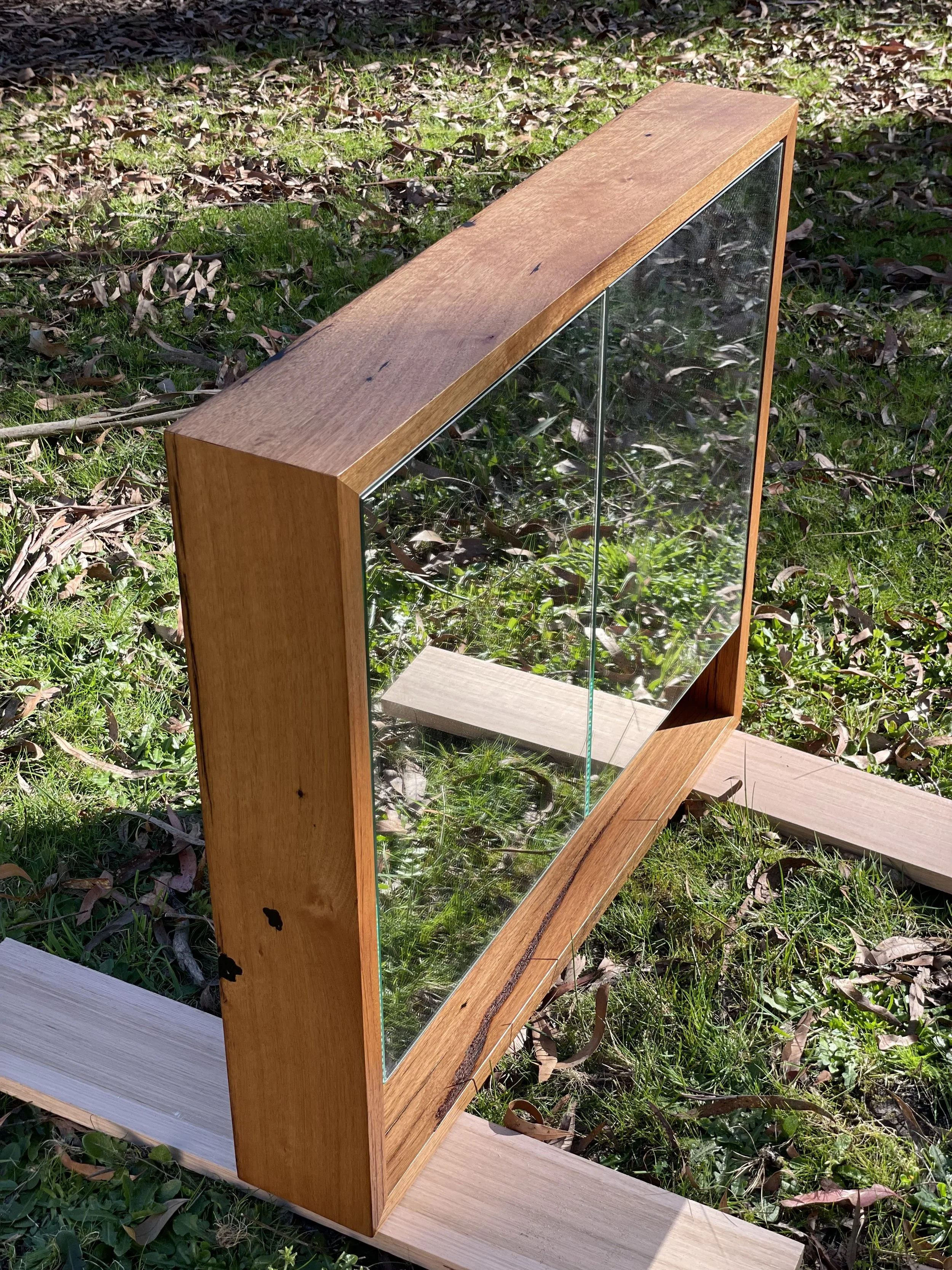 A wooden picture frame with glass, positioned outdoors on grass and leaves, leaning on a wooden plank.