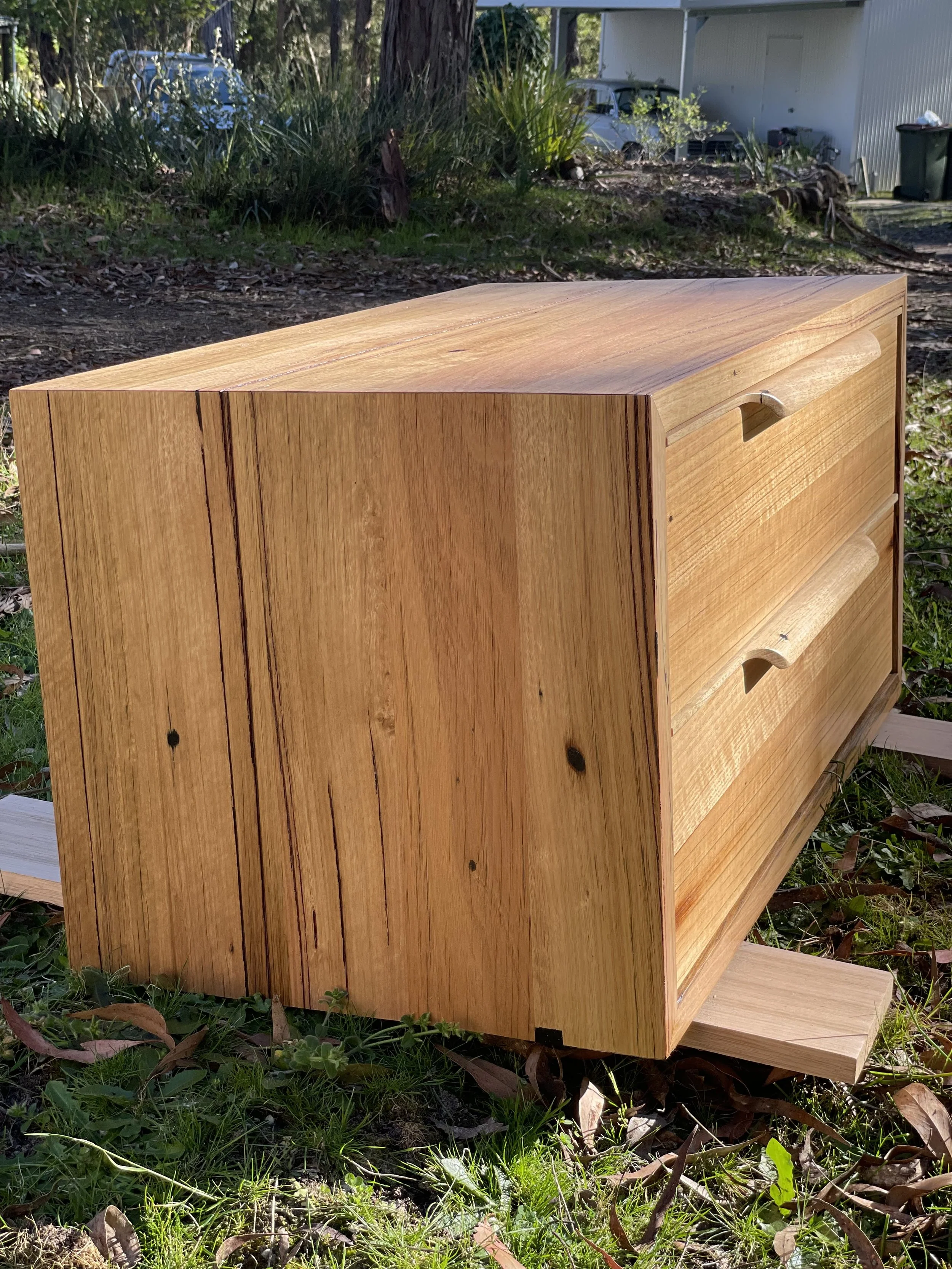 A wooden dresser with two carved handles on the front, placed outdoors on grass and leaves, with a background of trees, plants, and a building.