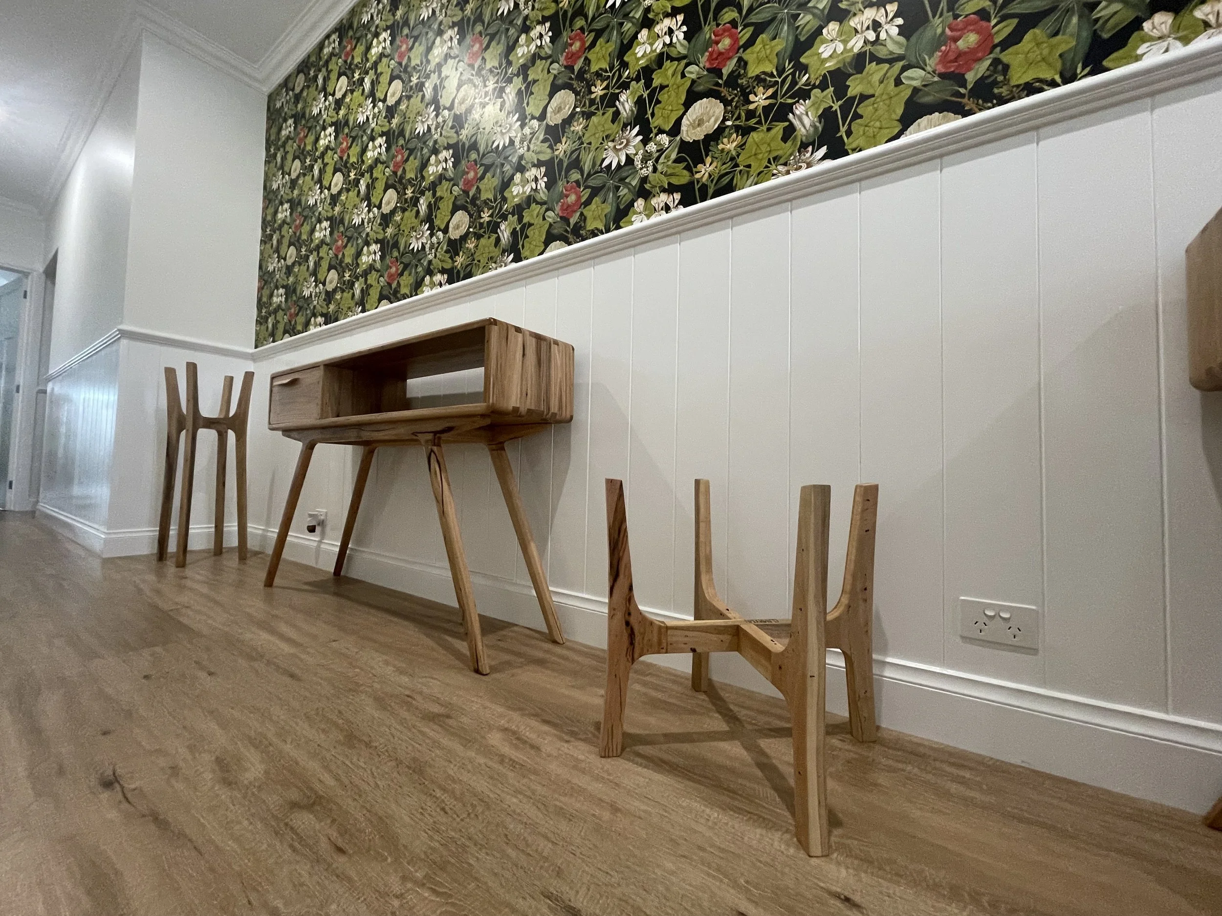 Wooden furniture including a small chair, a small table, and a desk, set against a white wainscoting wall with a floral wallpaper above.