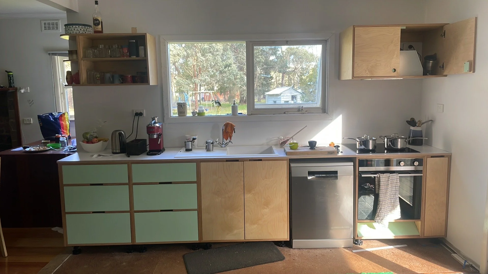 Kitchen with window, green and wood cabinets, countertop appliances, pots on stove, and a window view of trees and a backyard.
