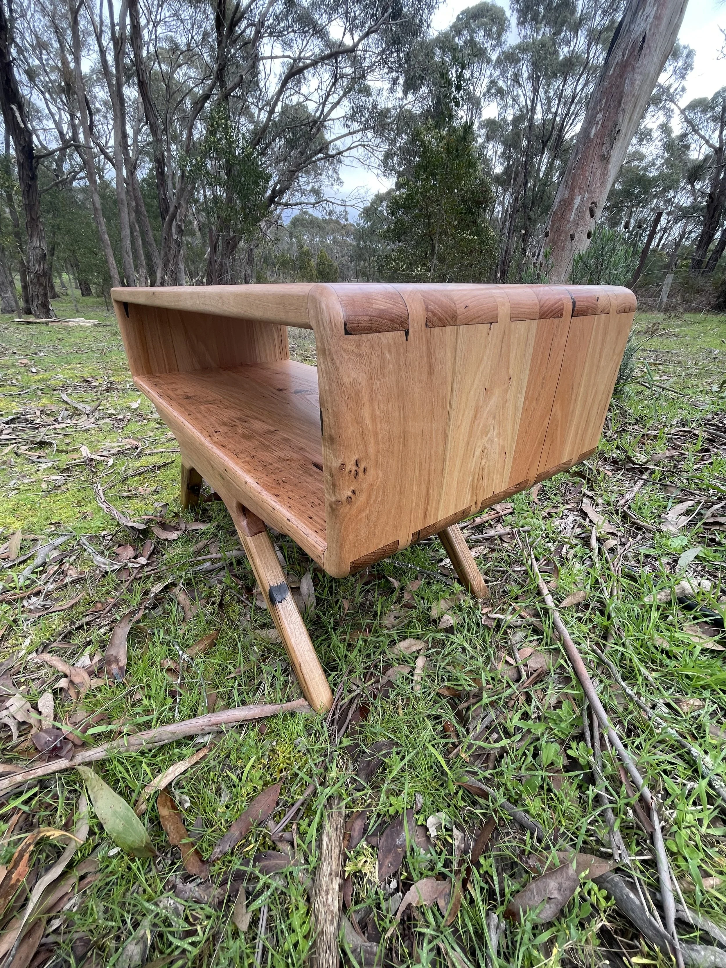 A wooden bench with a high backrests and angled legs sits outdoors on green grass and fallen leaves, with trees and an overcast sky in the background.