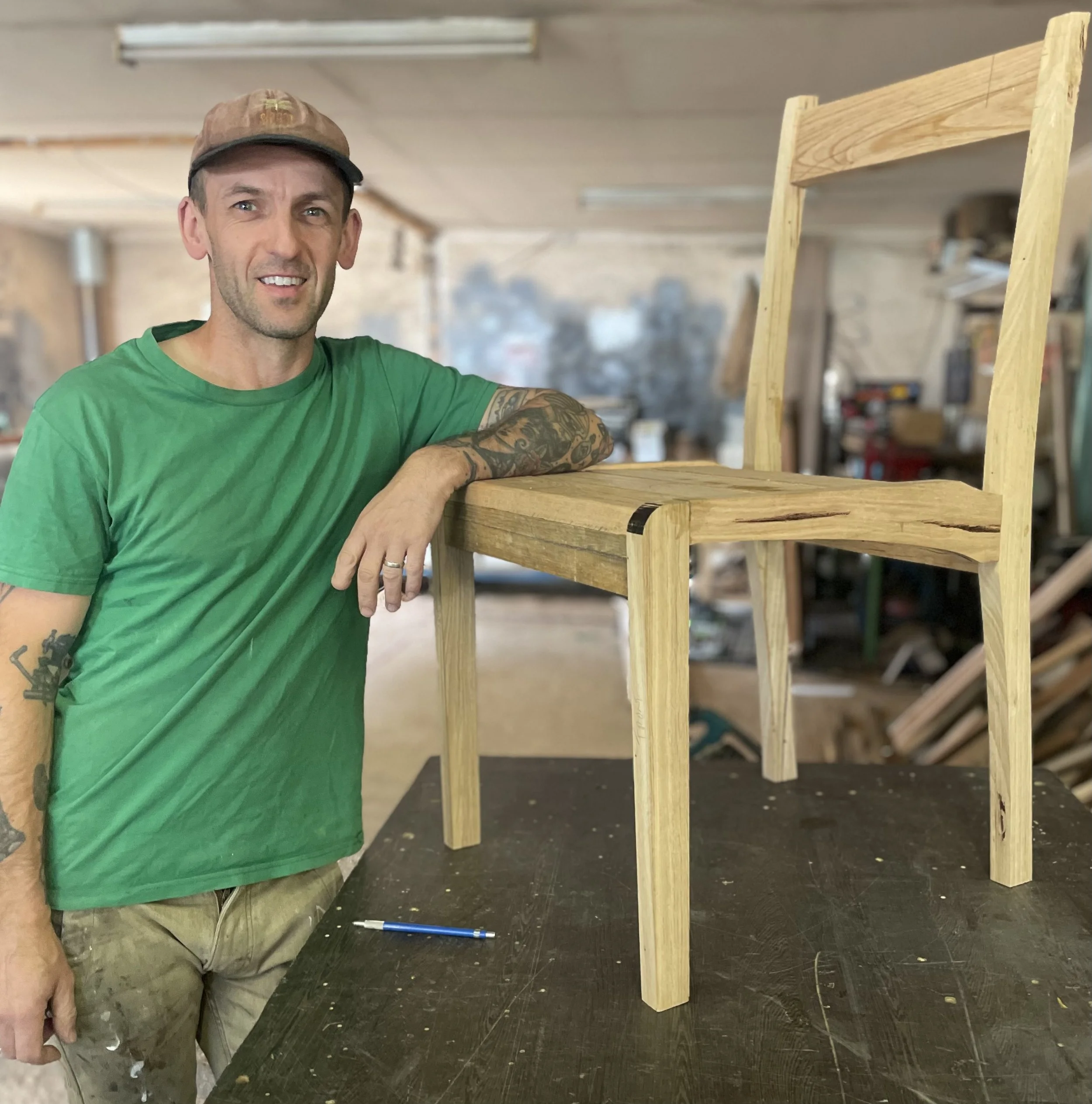 A man wearing a green t-shirt and a brown cap stands in a woodworking shop, posing next to a wooden chair frame on a work table.