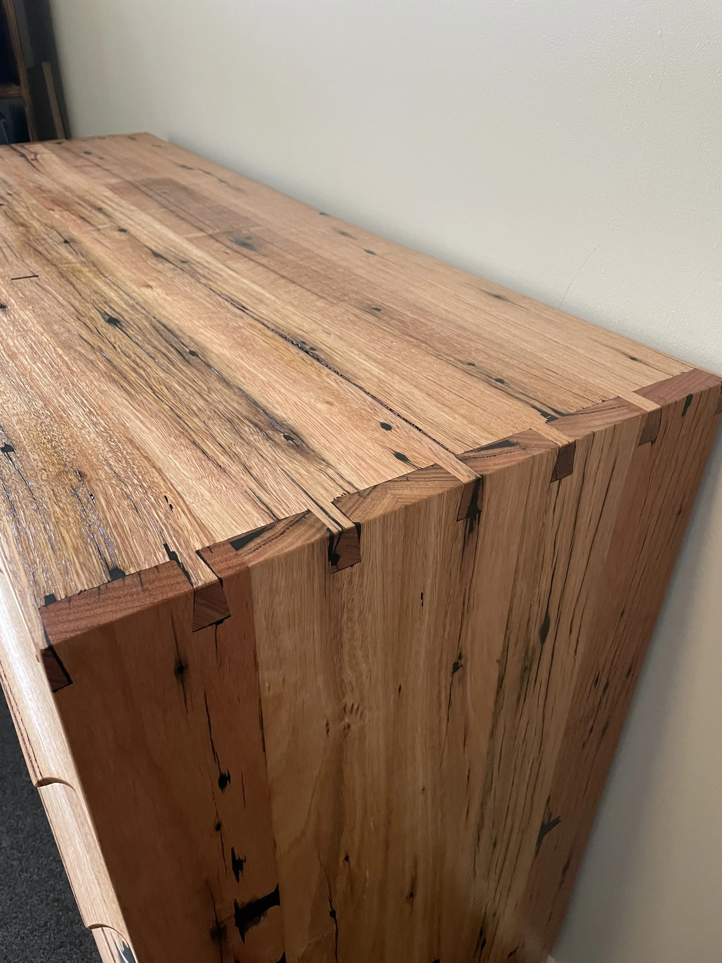 Close-up view of a wooden furniture piece with dovetail joints and a natural finish, placed against a plain wall.