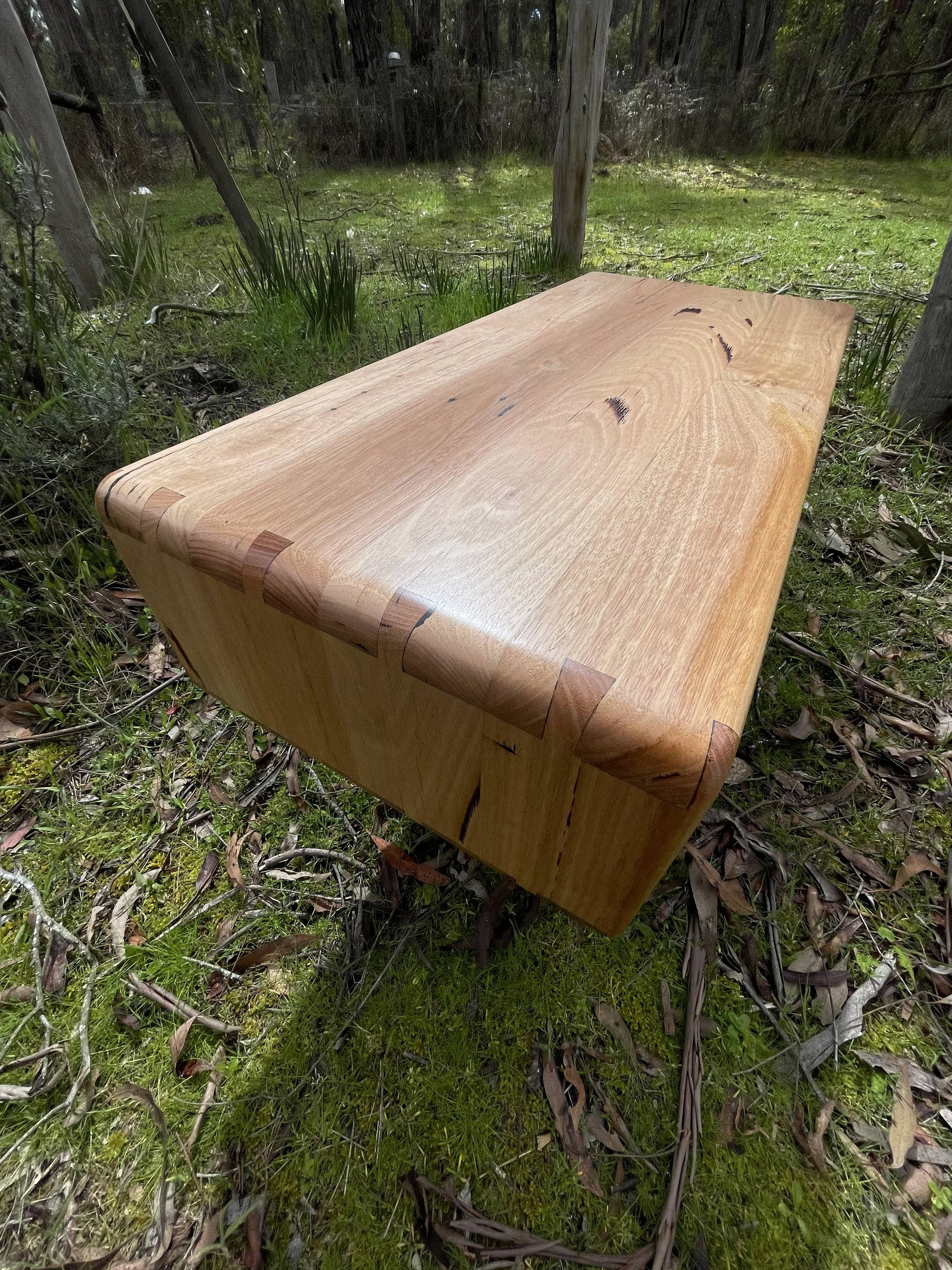 A wooden table with dovetail joints on the edges is placed outdoors on grass and surrounded by trees and plants.