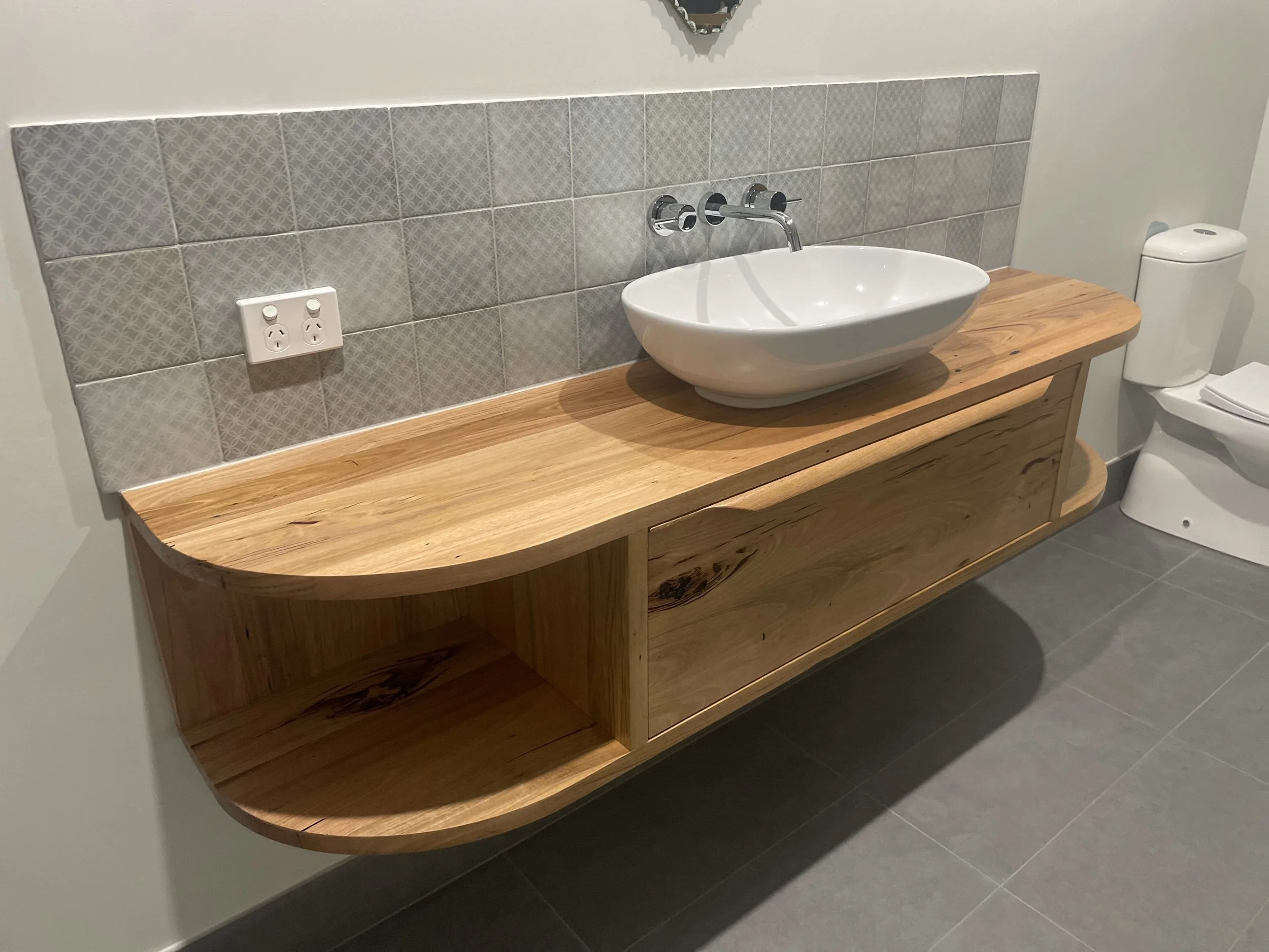 Bathroom vanity with a boat-shaped wooden countertop, a vessel sink, and a wall-mounted faucet. The wall behind the vanity has gray patterned tiles. There is a power outlet on the wall and a toilet to the right.