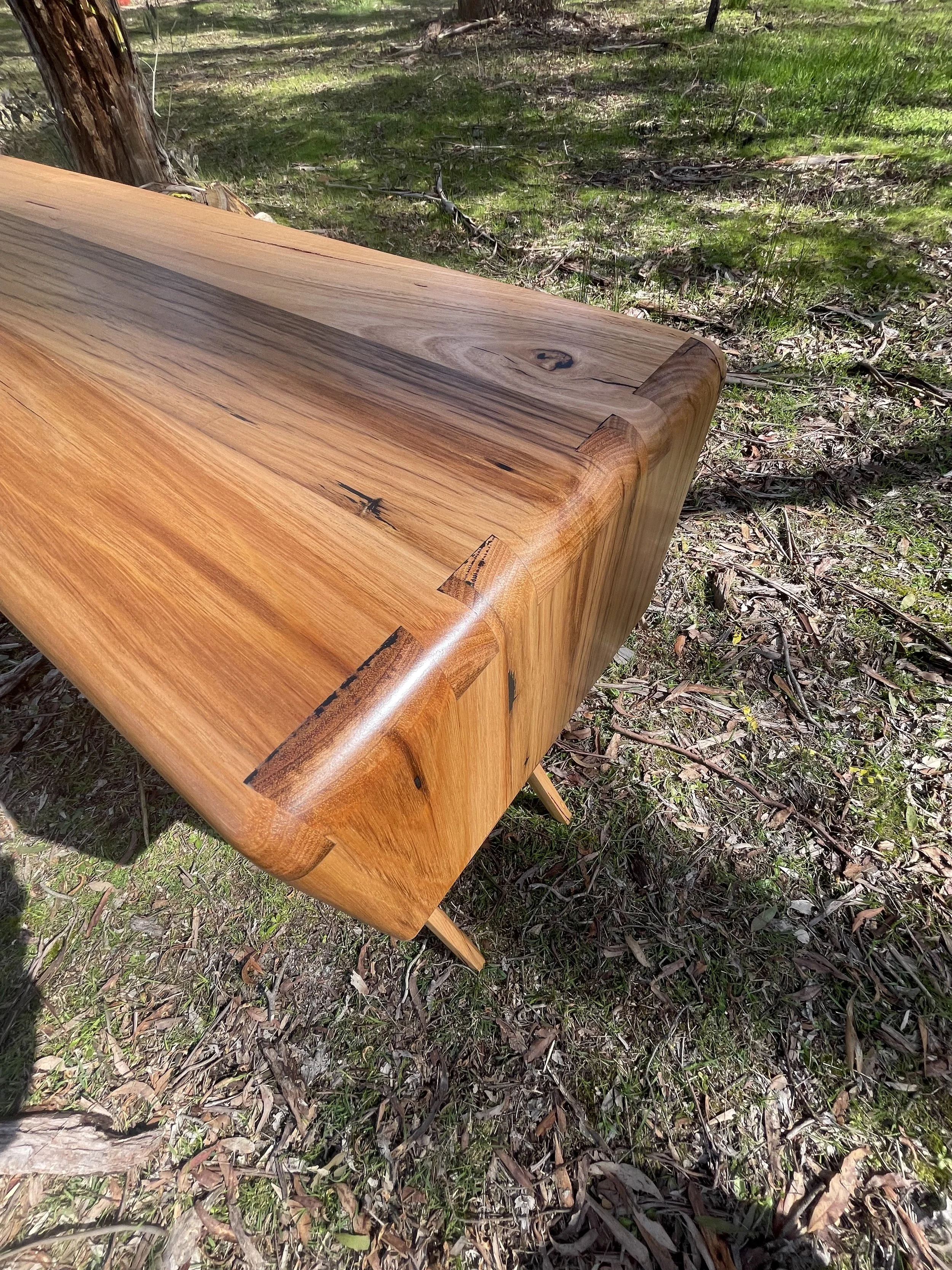 Close-up of a wooden table with a smooth, polished surface and rounded edges, set outdoors on grass and surrounded by trees.