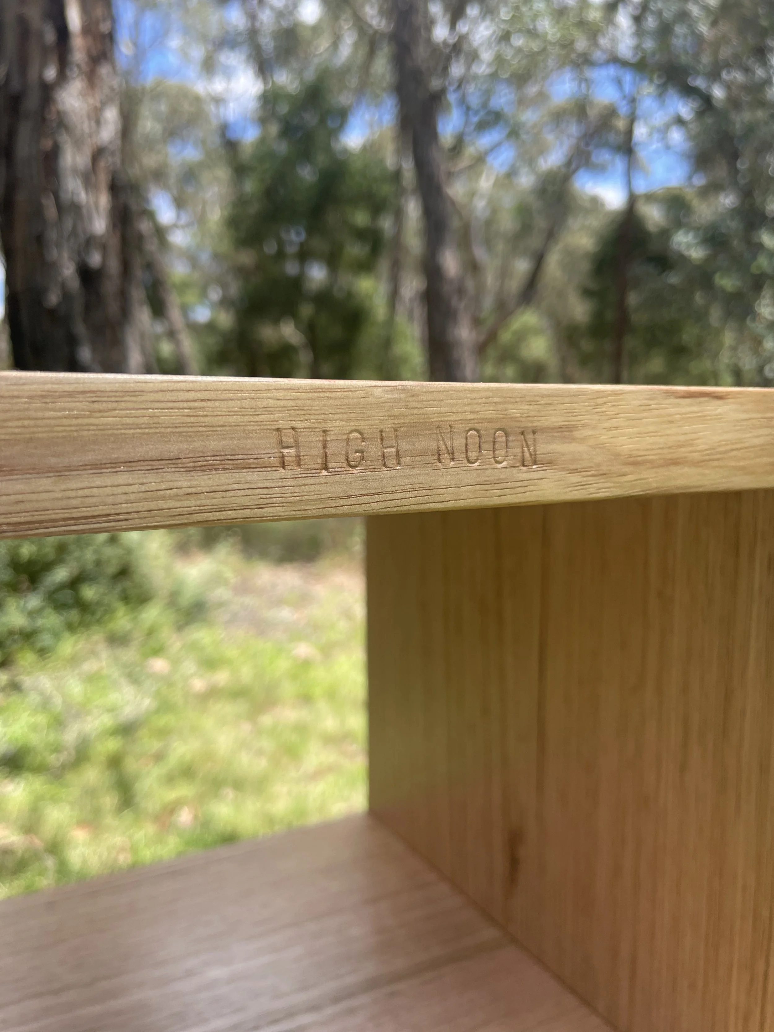 Close-up of a wooden piece with the words 'HIGH NOON' engraved on it, outdoors with trees and blue sky in the background.