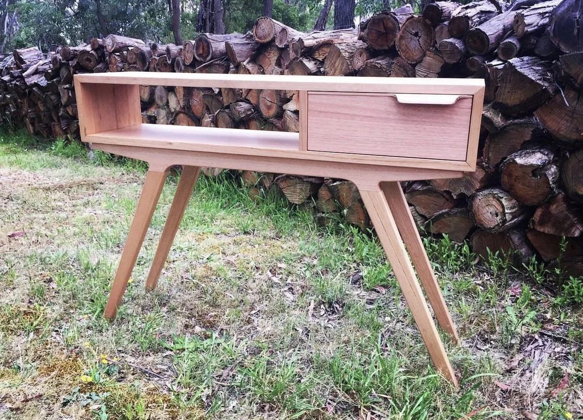 A wooden writing desk with a single drawer, set outdoors on grass, with a stacked pile of firewood behind it.