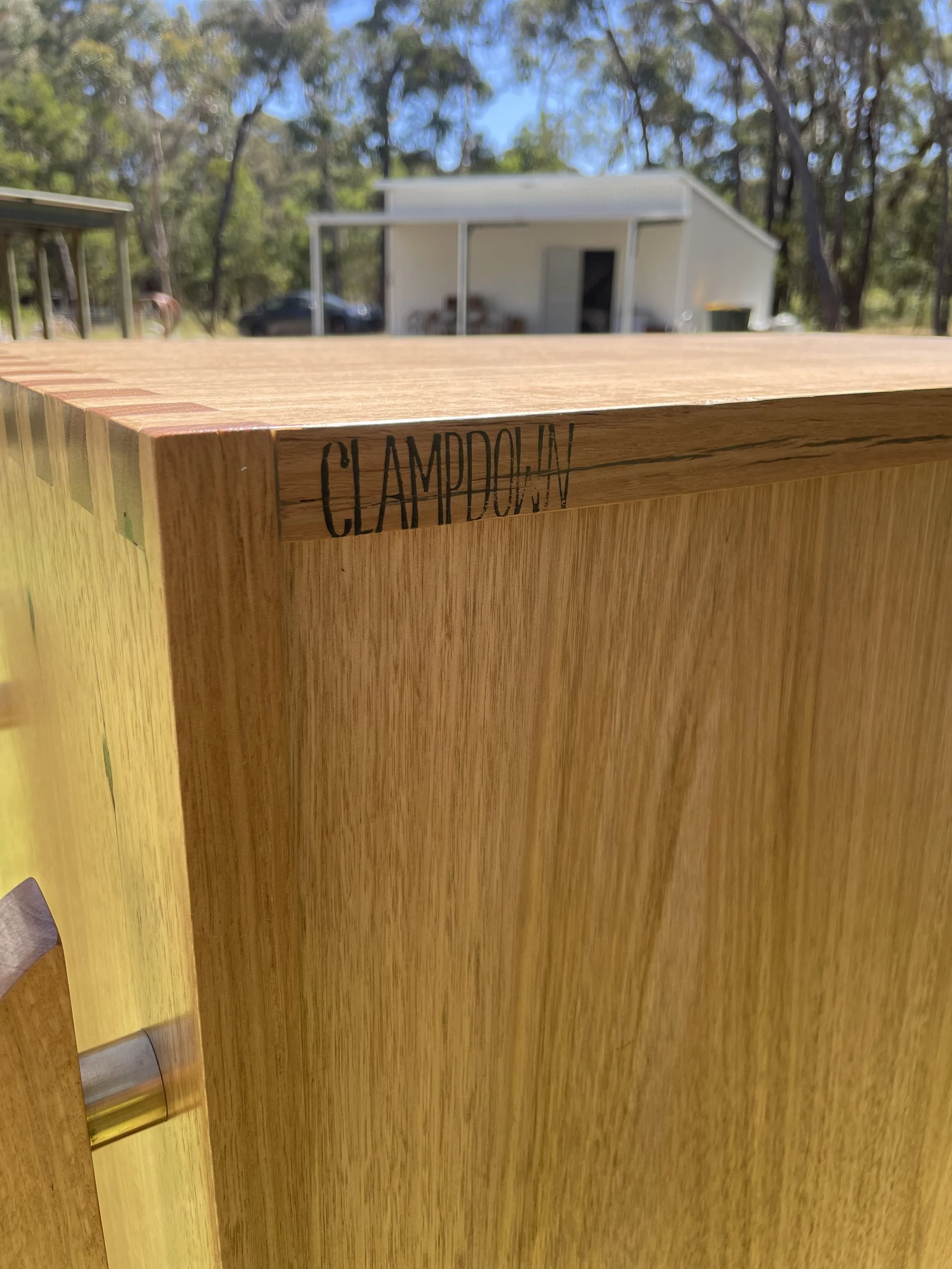 Close-up of a wooden cabinet showing the engraved name 'CLAMDOORWAY' on the top edge, outdoors with trees, a garage, and a vehicle in the background.
